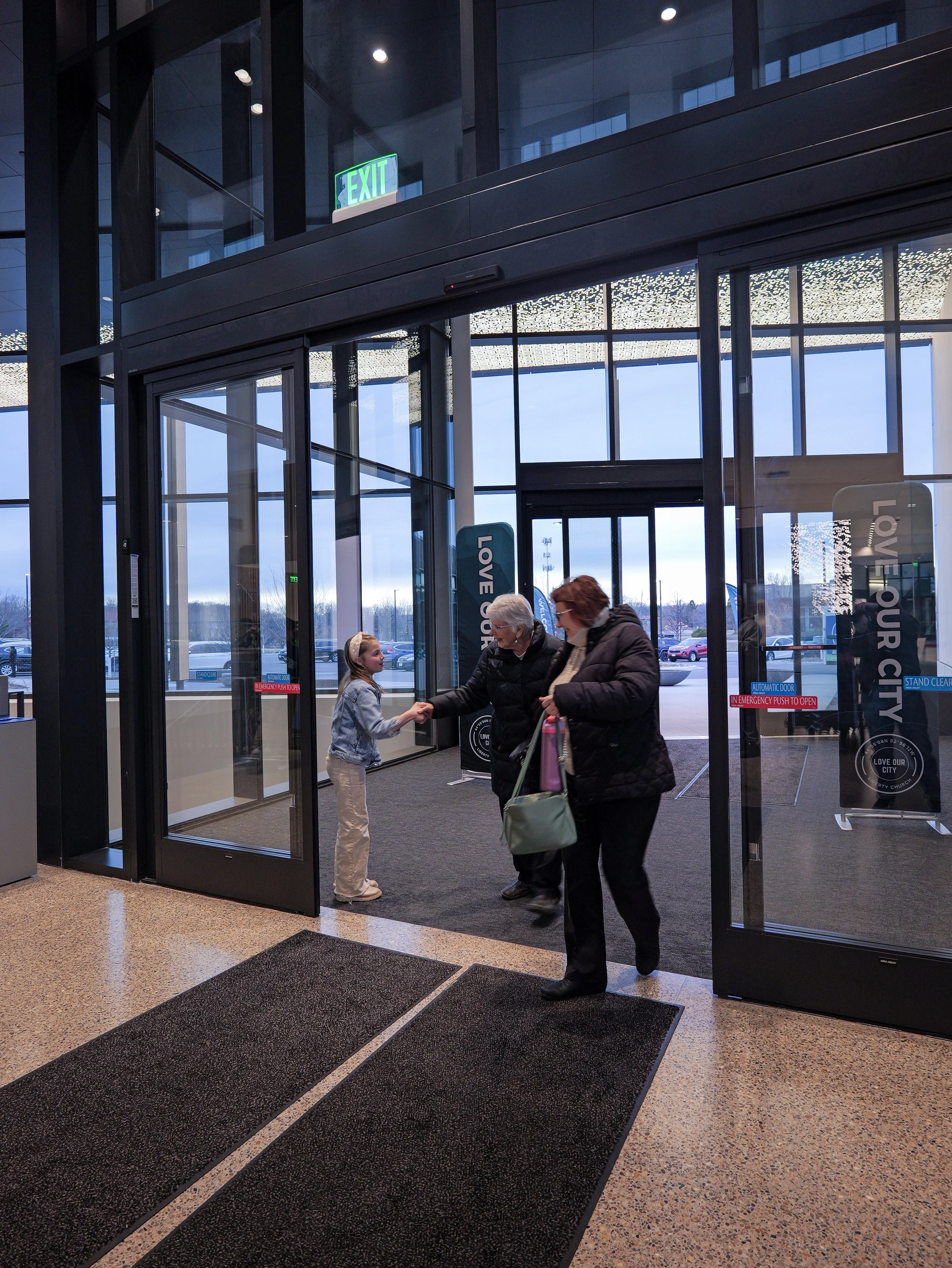 People entering through automatic glass doors at a spirit filled church in Woodbury, with an exit sign visible above. Inner lighting and exterior parking lot are visible.