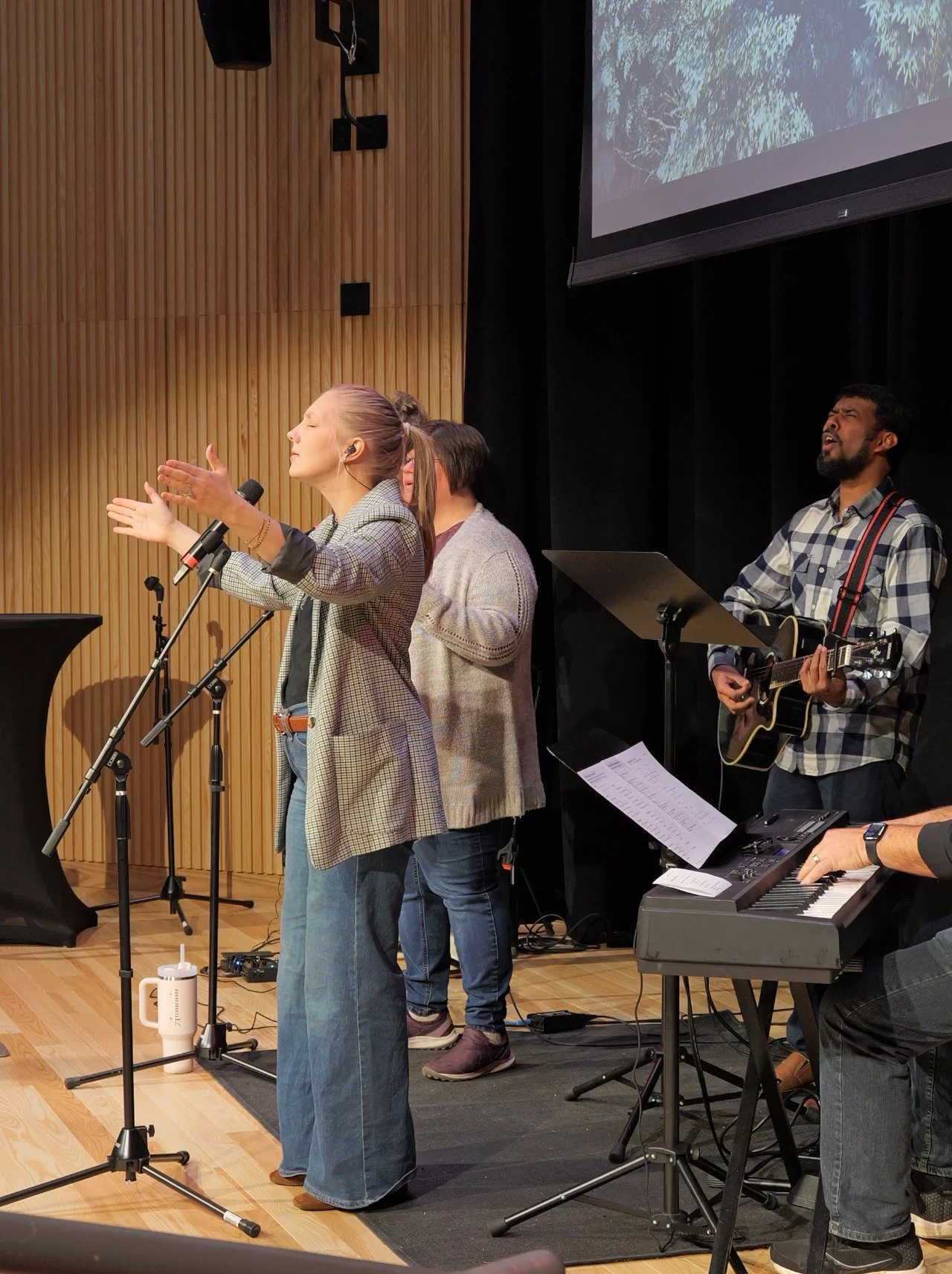 A group of people performing music on stage: a woman with a microphone singing with closed eyes, a man playing guitar, and a person playing keyboard at a modern worship church in Woodbury.