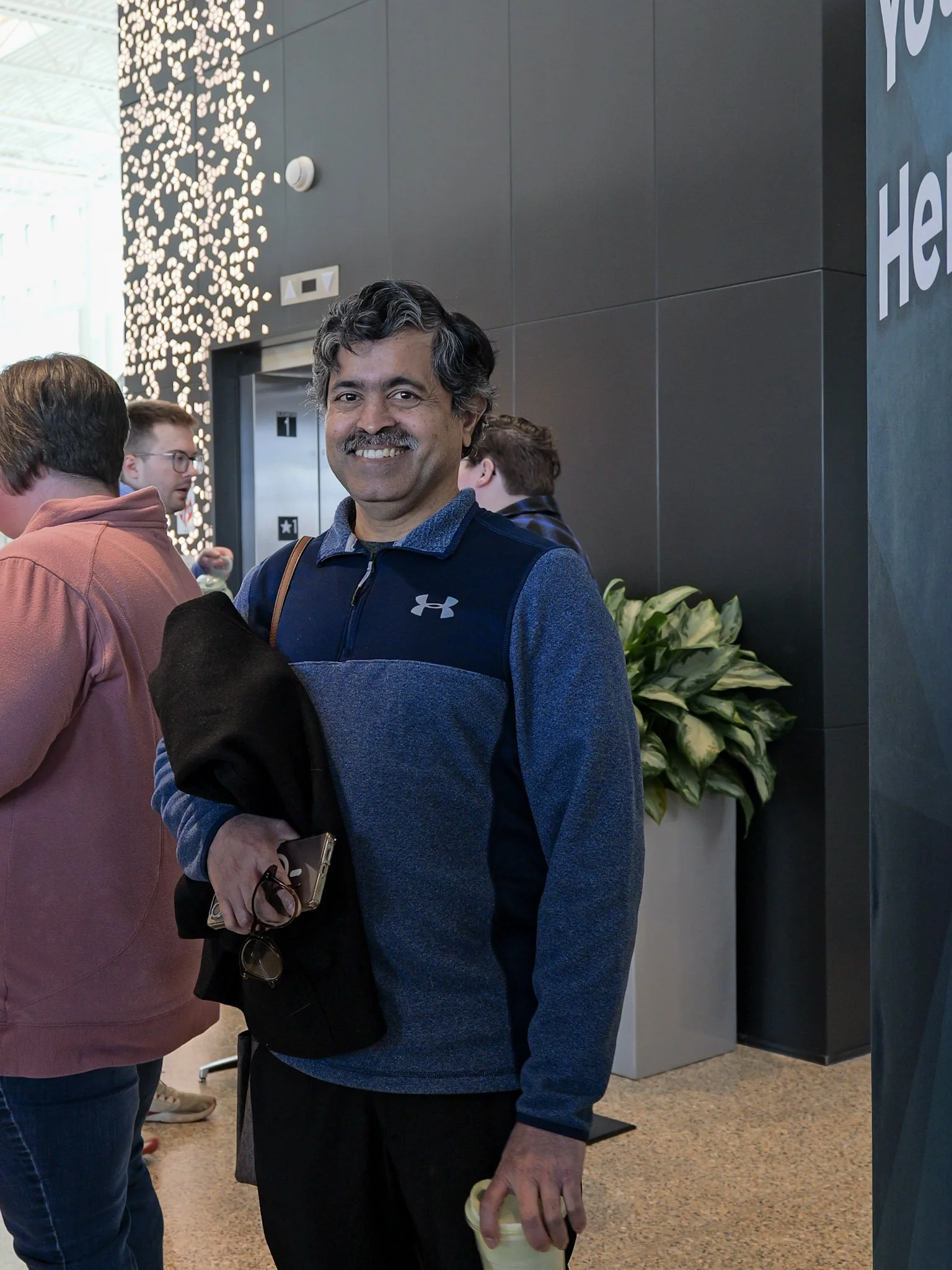 Smiling man in a blue jacket holding a cold drink, standing in a lobby with dark and gray walls, a plant, and a decorative light installation.