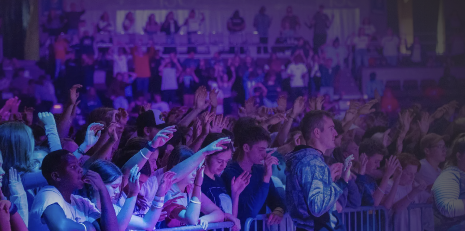 Crowd of youth at a church event, some with raised hands, in a room illuminated with colorful lights.
