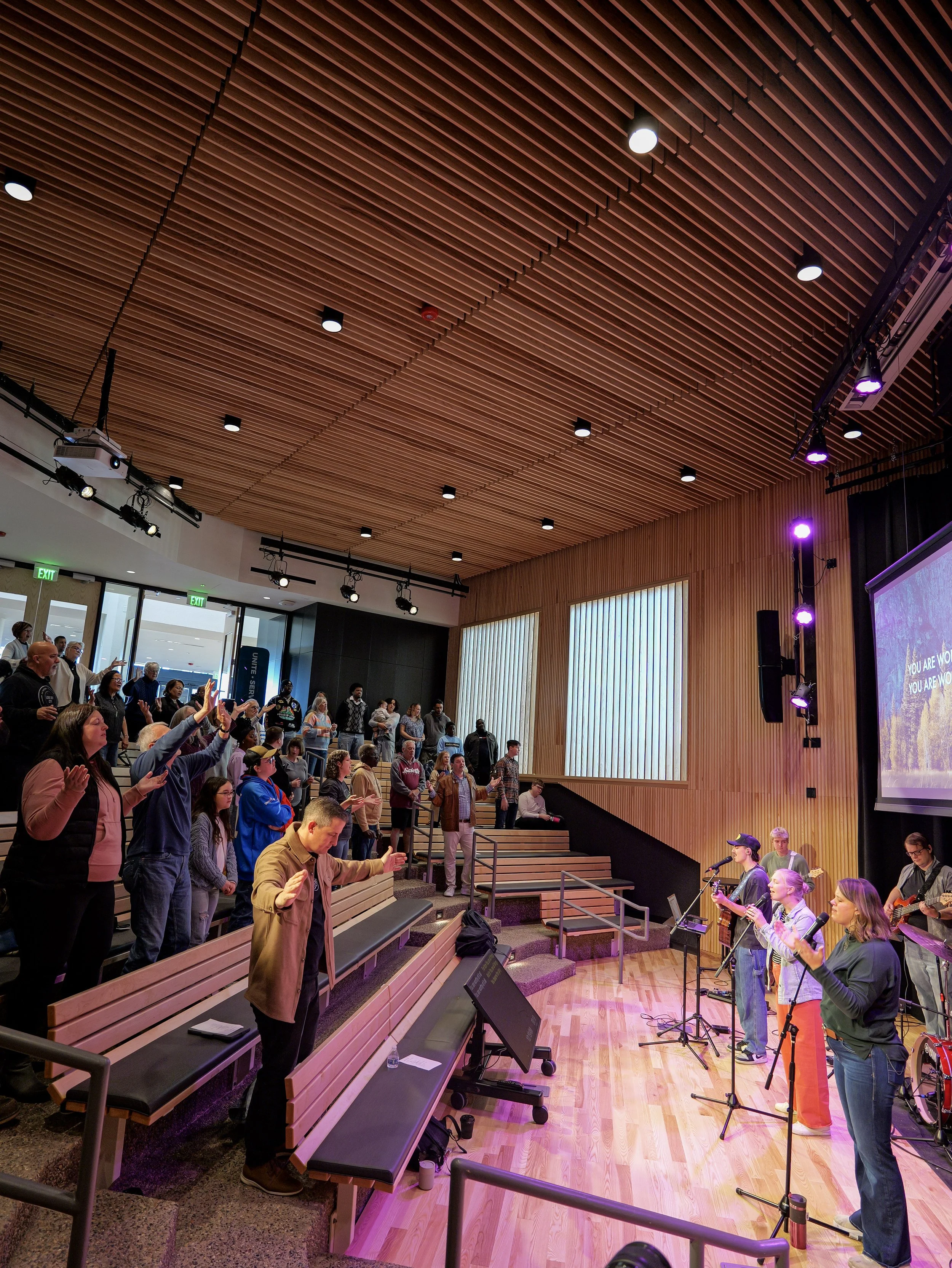 People gathered in a modern auditorium with wooden walls and ceiling, and engaging in a worship service. Some audience members are standing with hands raised, as musical instruments are played