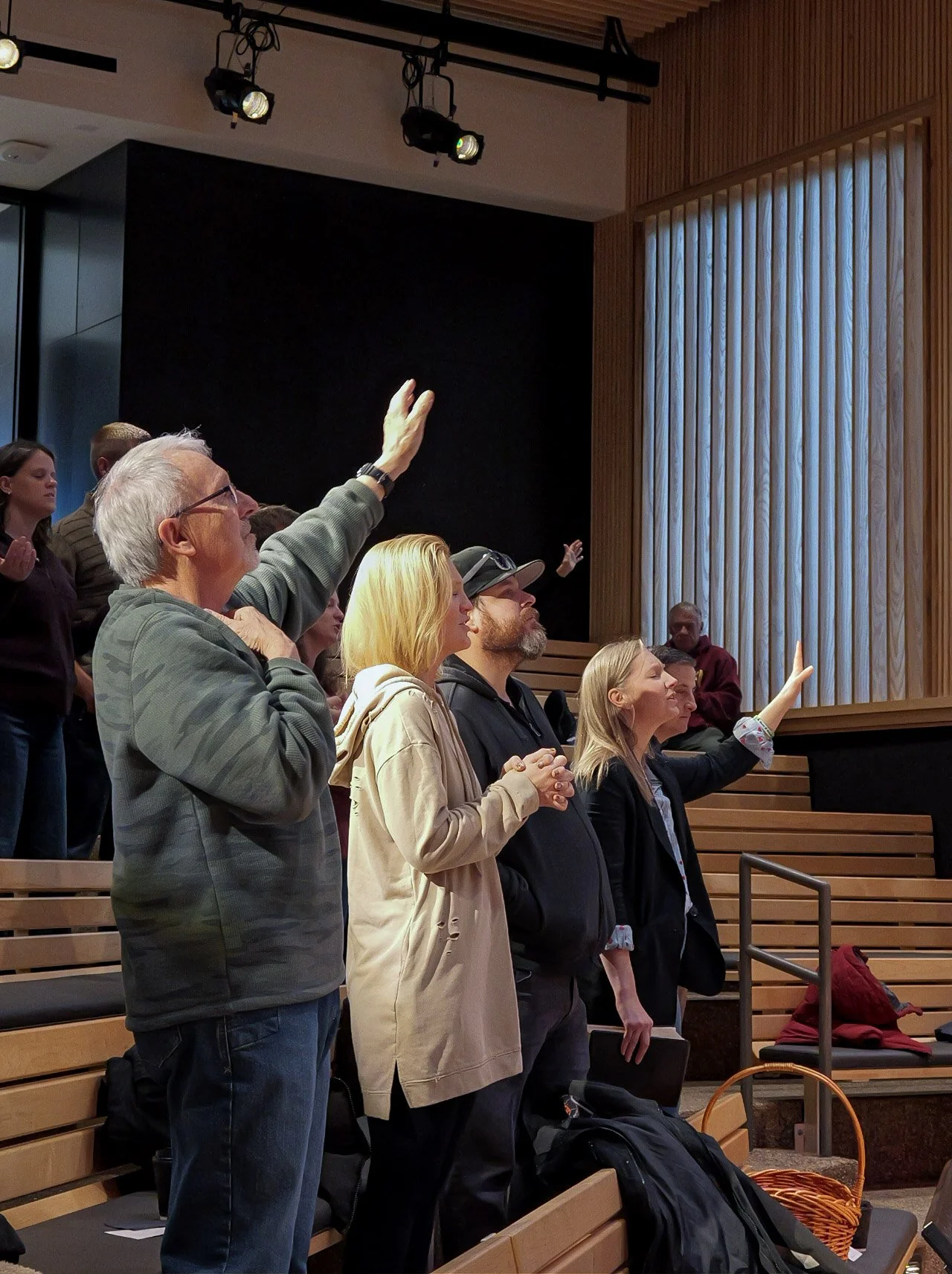 Group of people, both men and women, raising their hands in a meeting or conference room with wooden walls and seating.