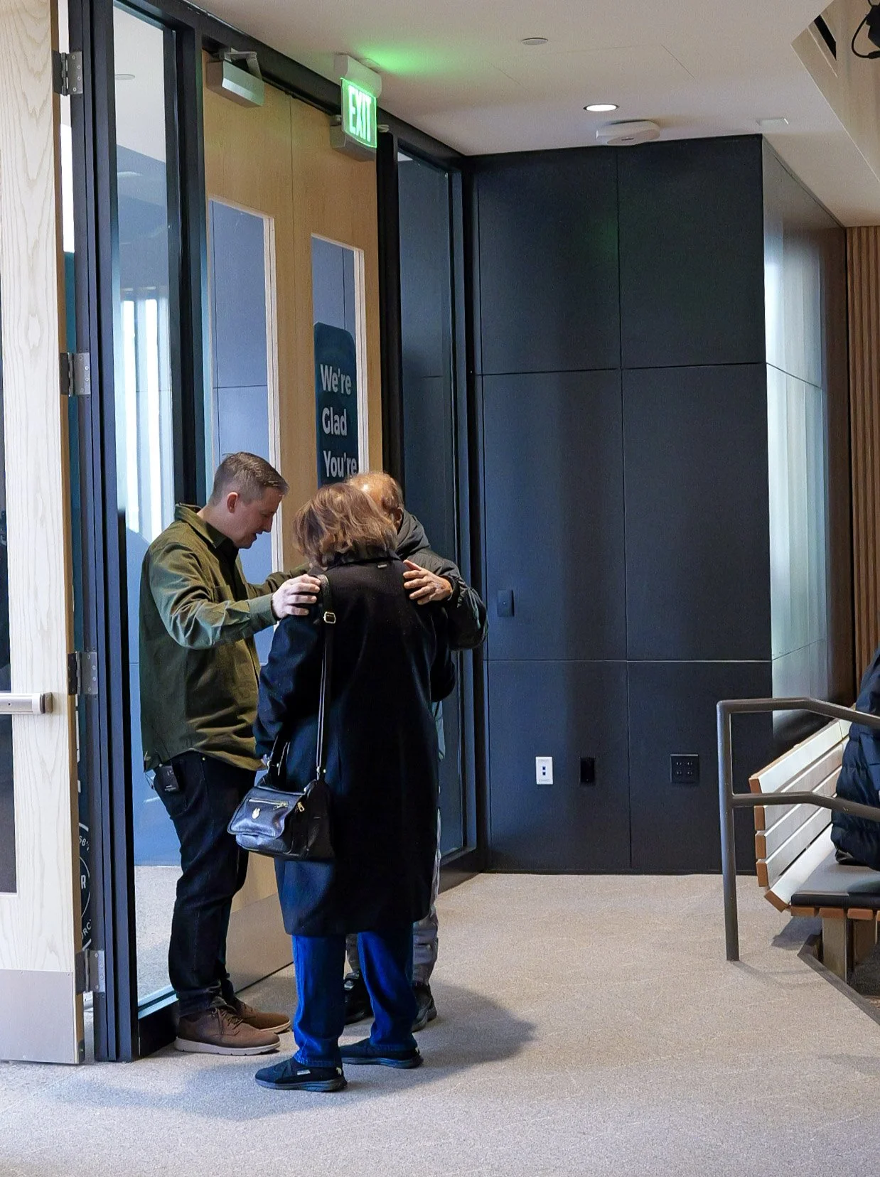 Three people praying together in a hallway at a spirit-filled church in the twin cities area. The door has a sign that says, "We're Glad You're".