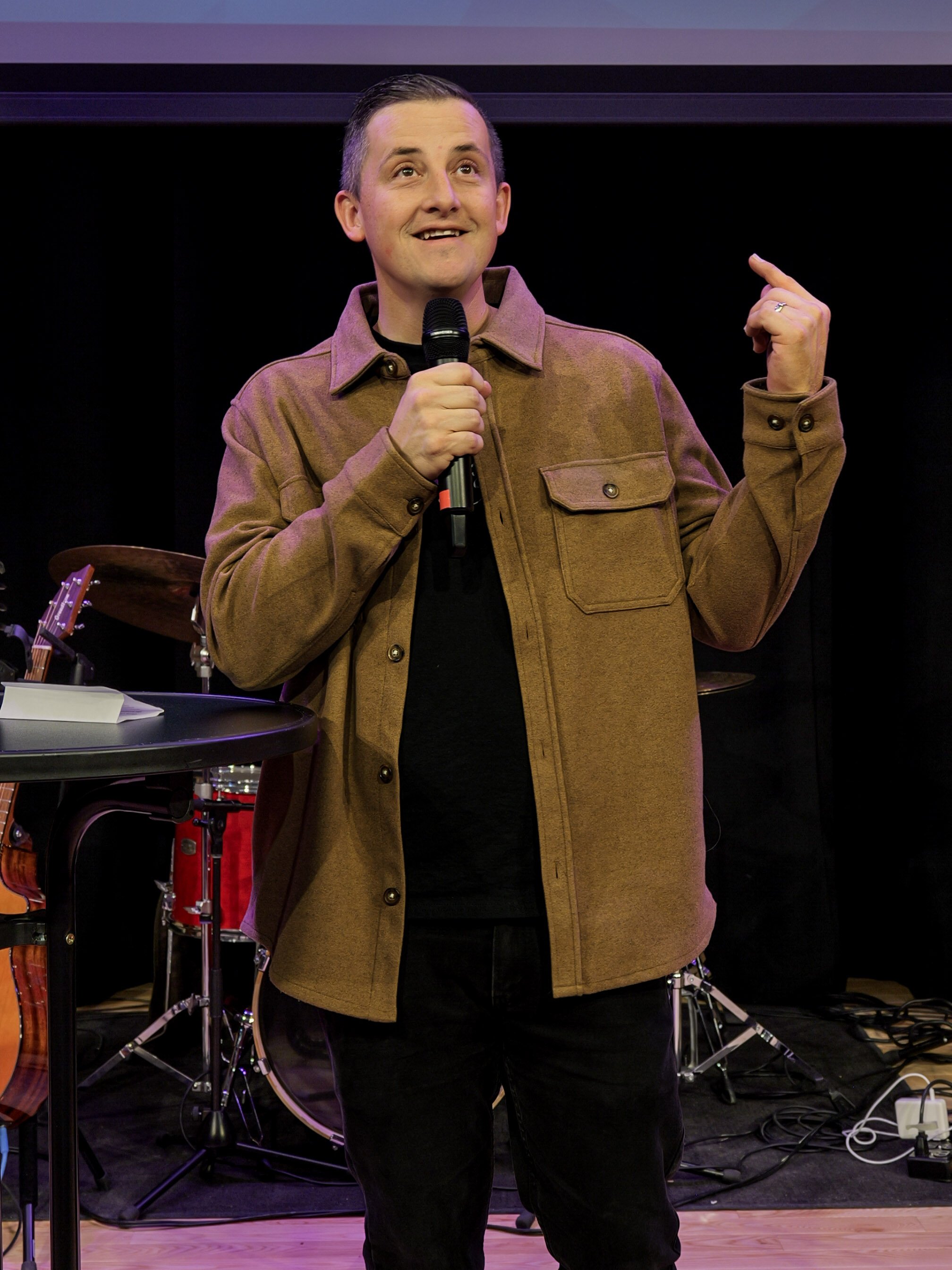 A pastor of a welcoming Woodbury Minnesota church holding a microphone, gesturing with his right hand, standing in front of musical instruments and equipment on a stage.