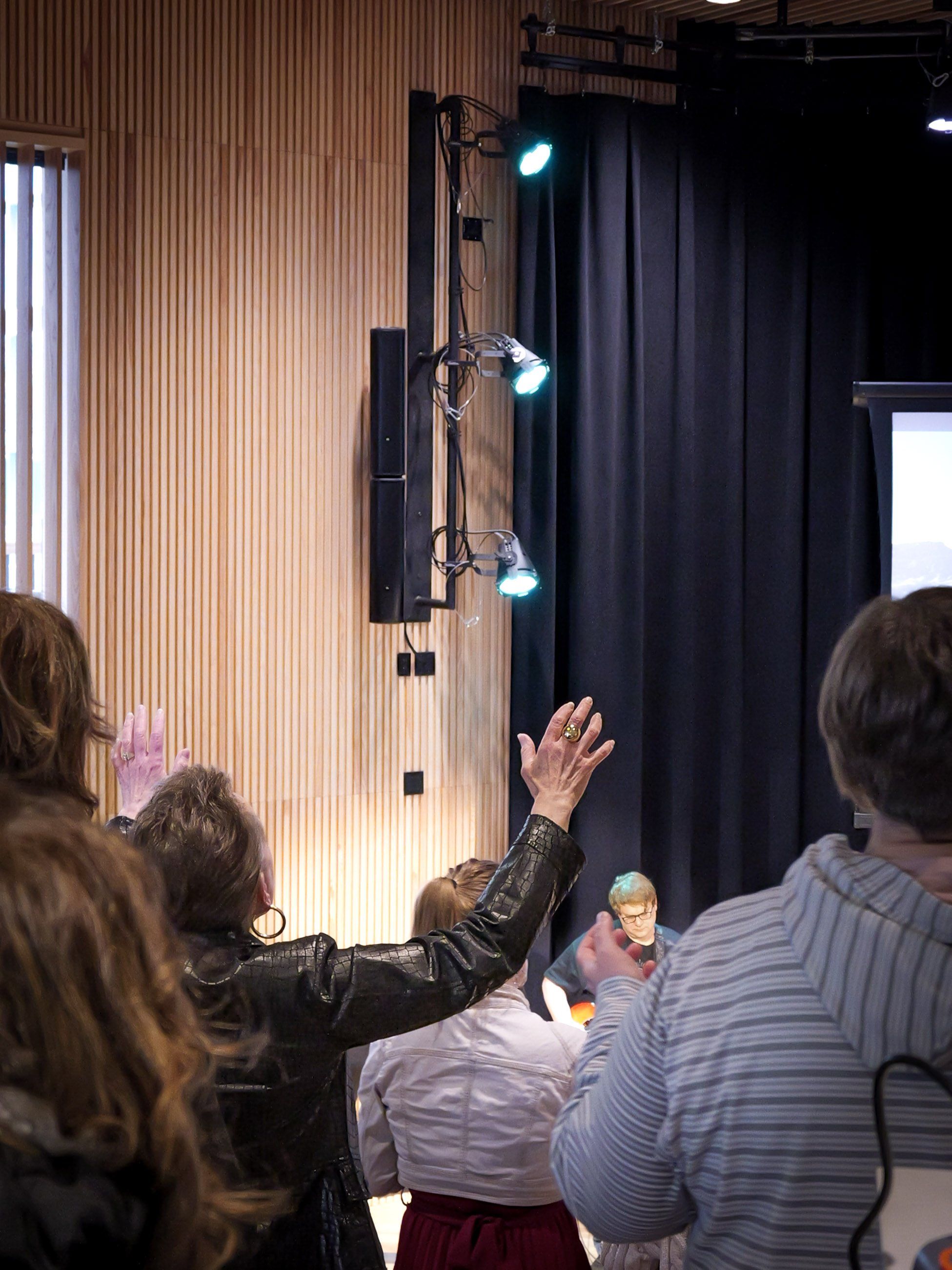 Audience raising hands during a worship service at a Spirit filled church in Woodbury in a room with wood-paneled and black curtain walls.