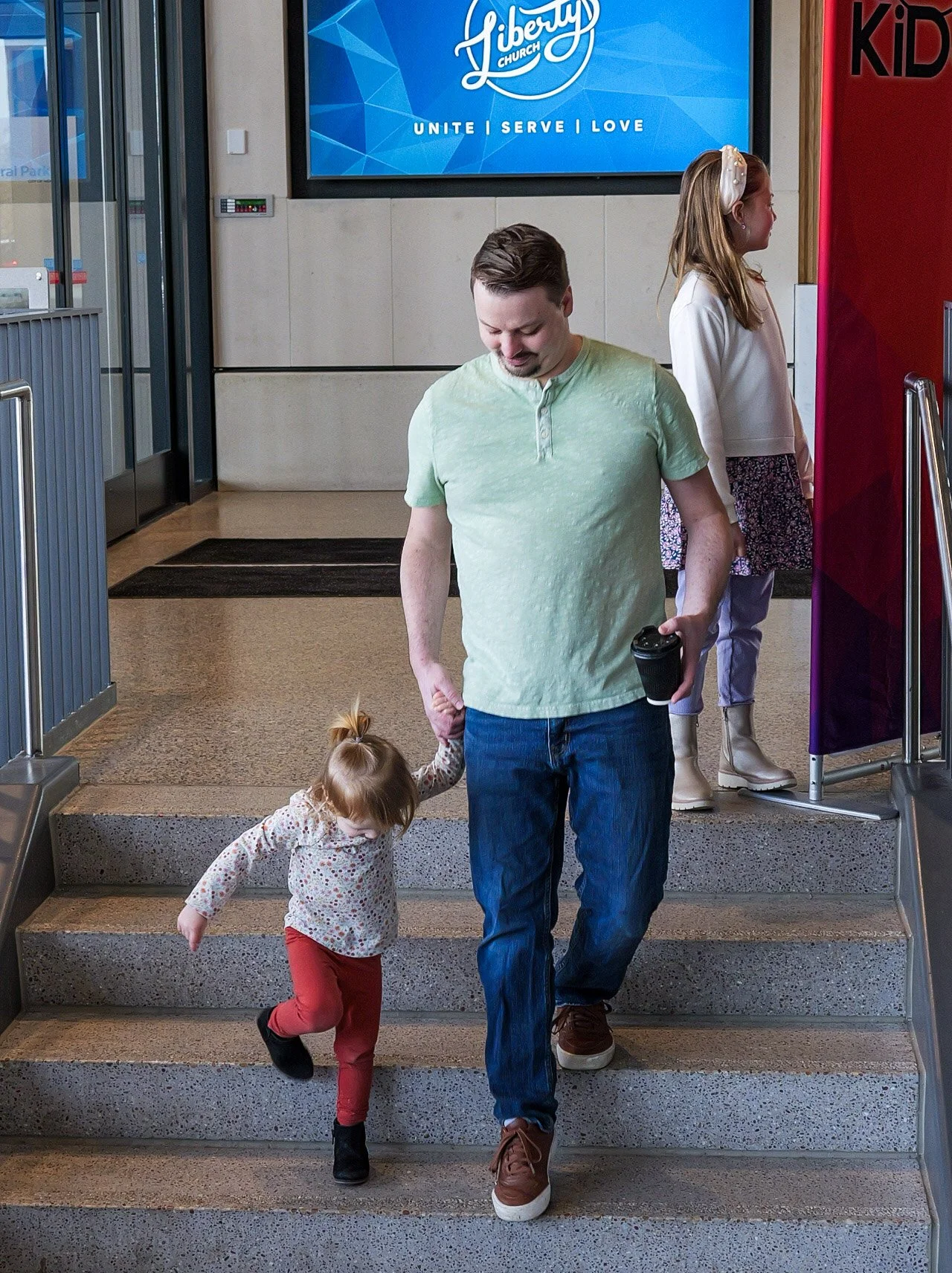 A man and two young girls walk down stairs in a family church Minnesota. The man is holding a coffee cup and a child's hand. There is a large blue sign with the word 'Liberty' and words 'Unite, Serve, Love' in the background.