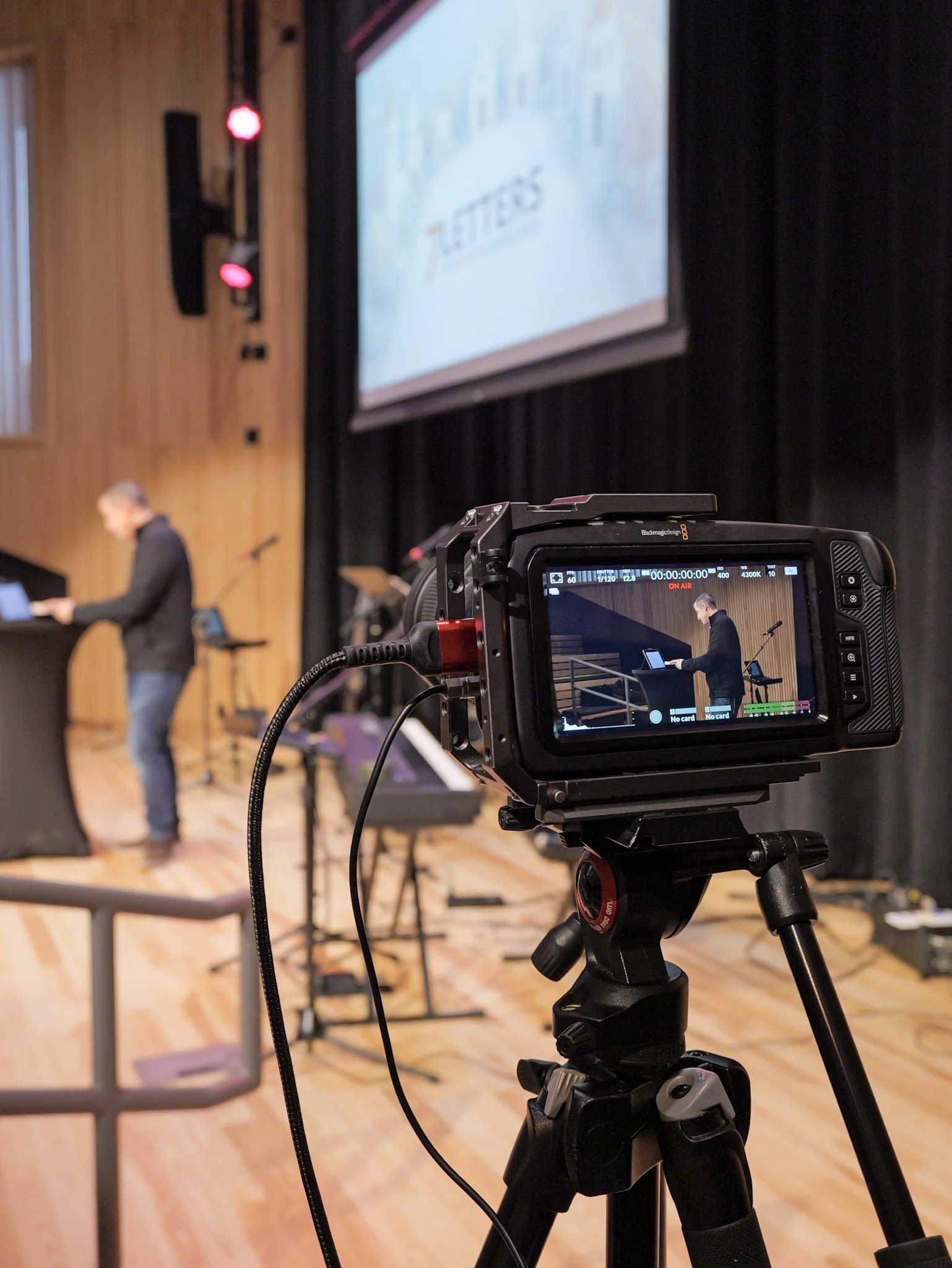 Camera capturing a man preaching a bible based message at a welcoming church in Woodbury, with a large screen behind showing a blurred presentation or slide, in a wooden-paneled auditorium setting.