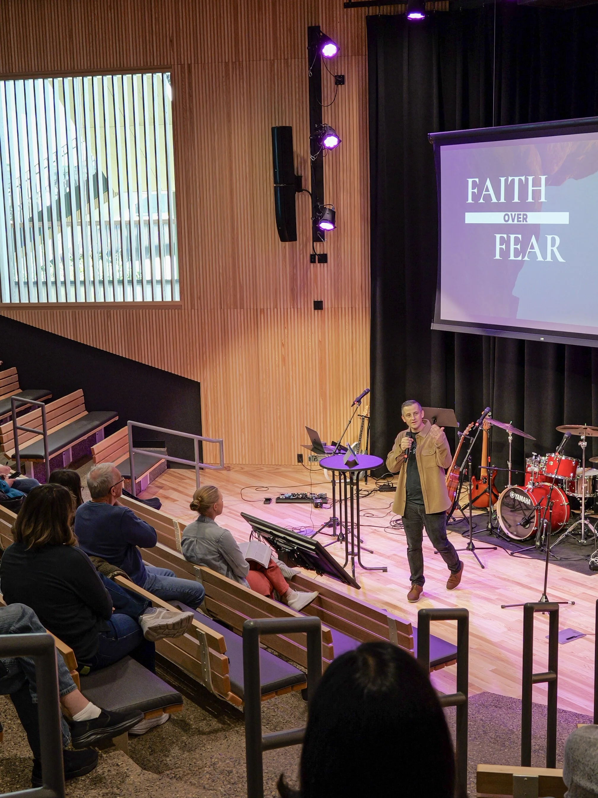A speaker in a brown jacket and black pants is giving a sermon on a stage in front of an audience seated in local church in Woodbury. A large screen displaying the words 'FAITH OVER FEAR.'