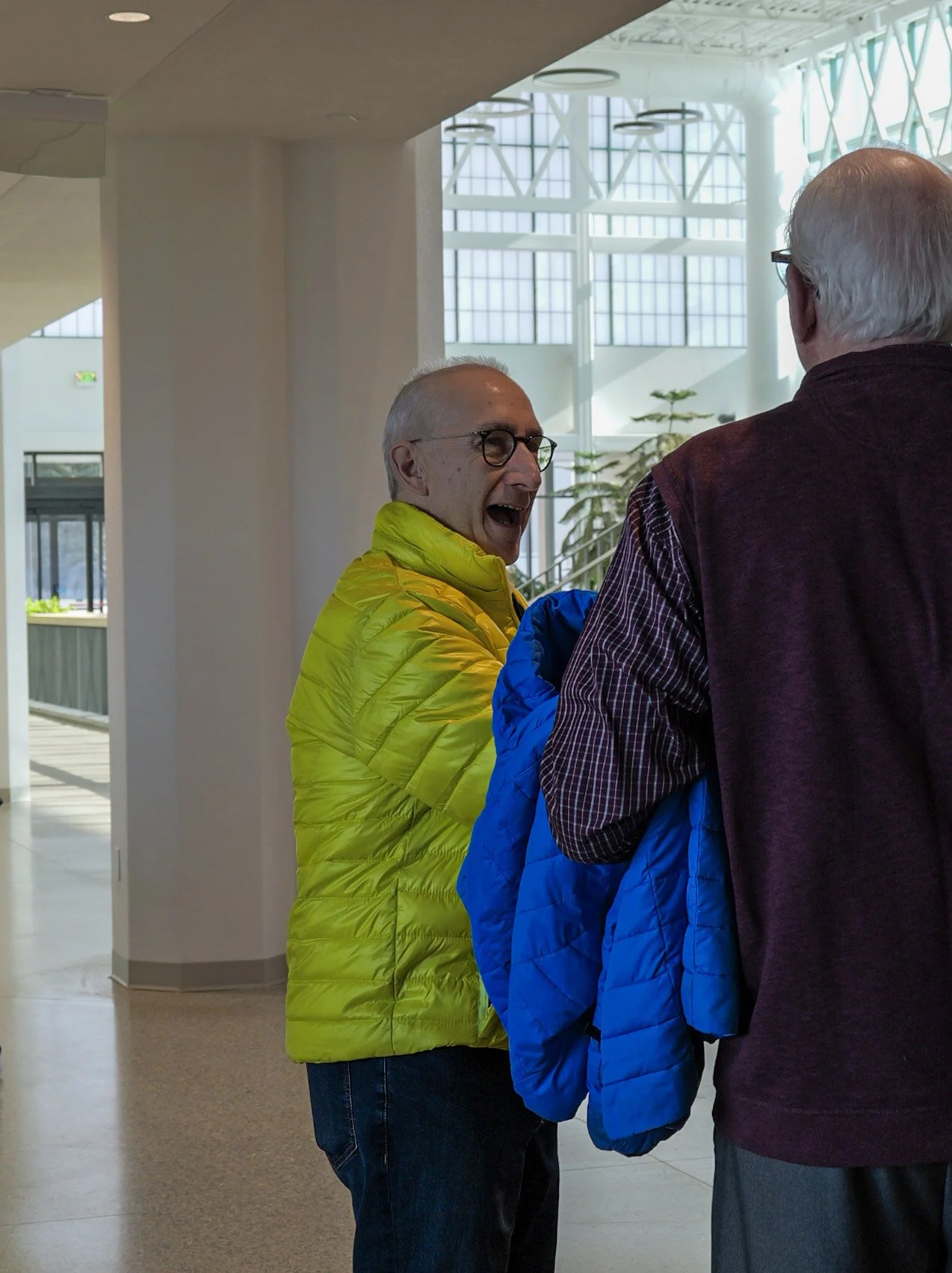 Two elderly men talking indoors, one wearing a bright yellow jacket and glasses, the other with gray hair and a checked shirt under a dark vest.