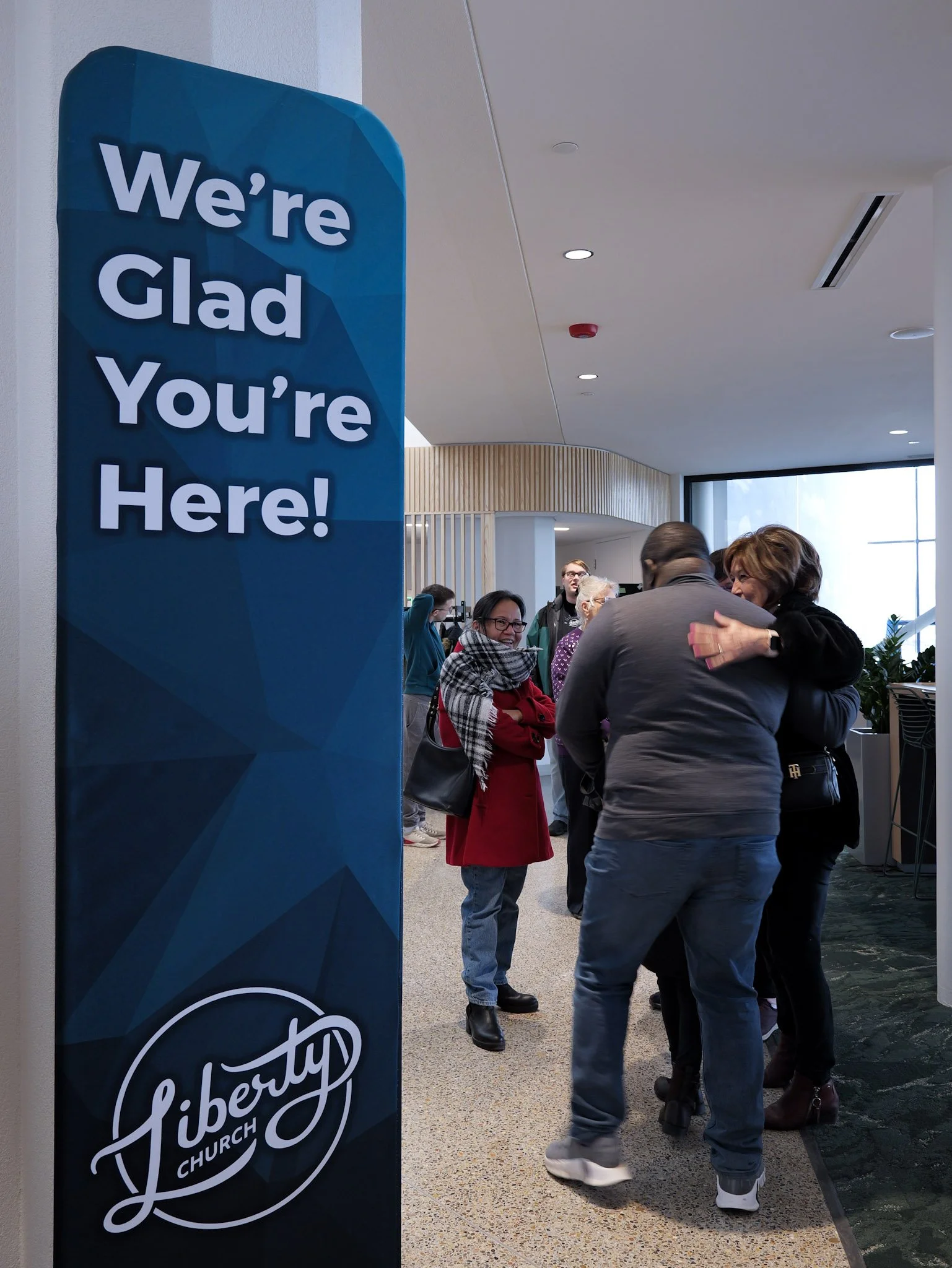 A welcoming and engaged diverse  group of people socializing indoors near a large window. A blue sign with white text reads, 'We're Glad You're Here!'.