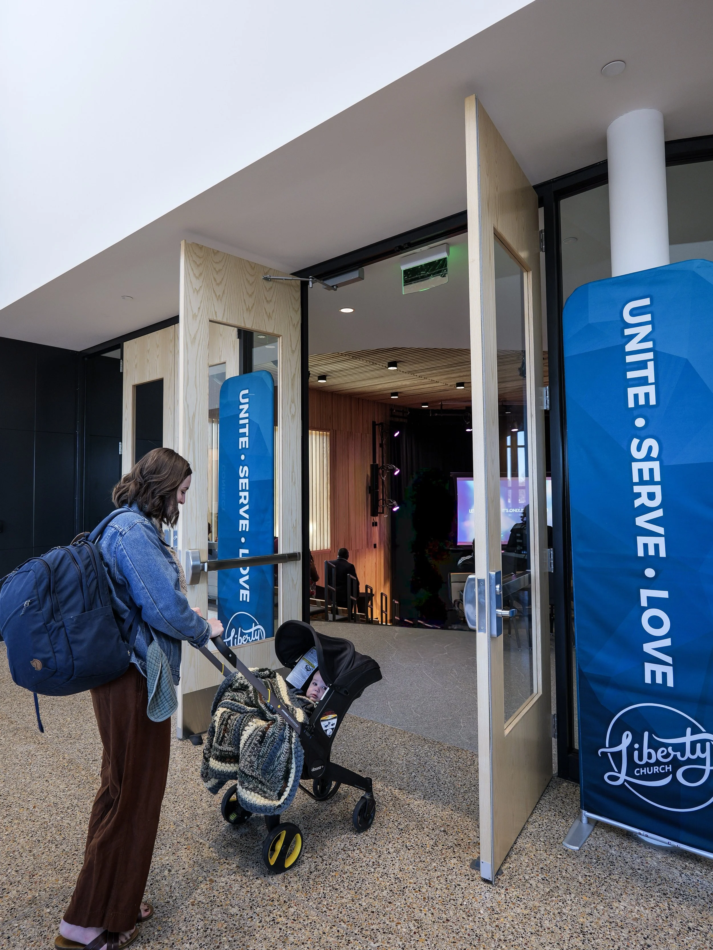 A woman standing outside Liberty Church, looking at a young child in a stroller. The entrance has a blue banner that reads “unite, serve, love.” A church for young families.