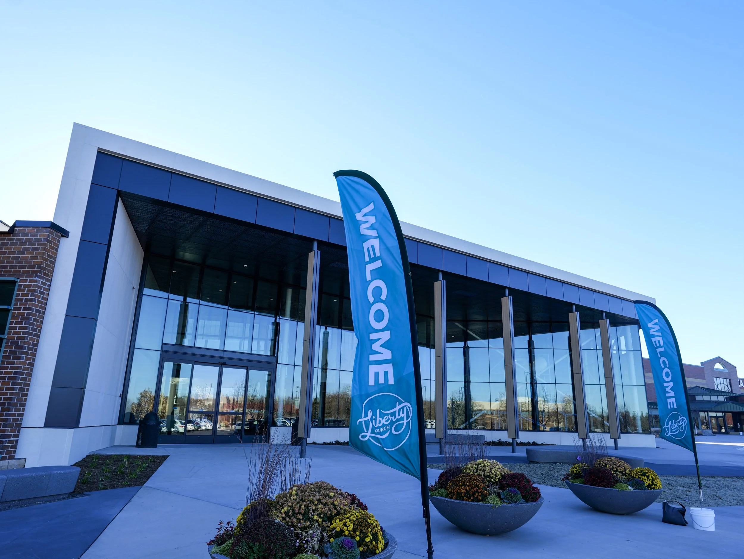 Exterior view of a modern church building with large glass windows. Two blue flags with white text reading 'Liberty Church' are positioned outside, with landscaped flower beds in large pots near the entrance. The sky is clear and blue.