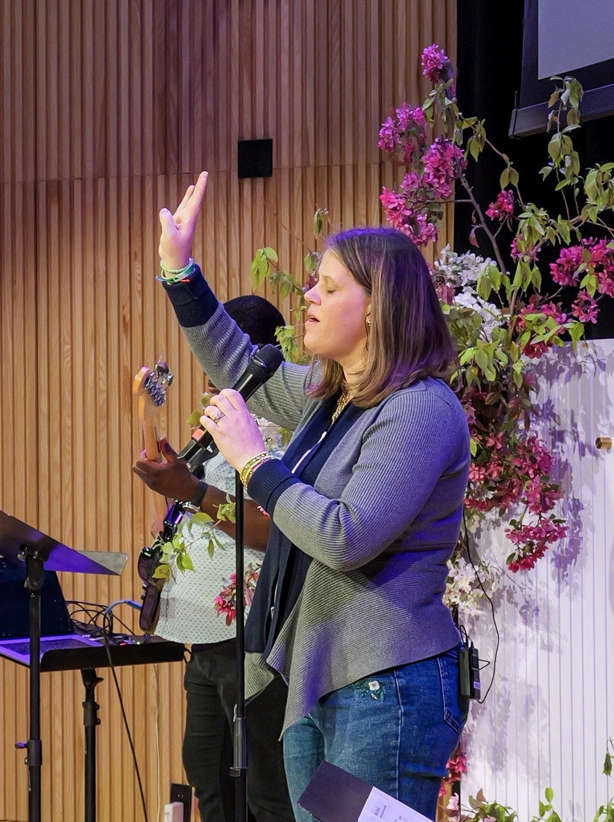 A woman with shoulder-length brown hair singing into a microphone with her eyes closed, raising one hand. She is wearing a gray lapel jacket over a dark top, and blue jeans. She stands in front of a floral arrangement and a wooden panel wall, with a person playing guitar in the background.