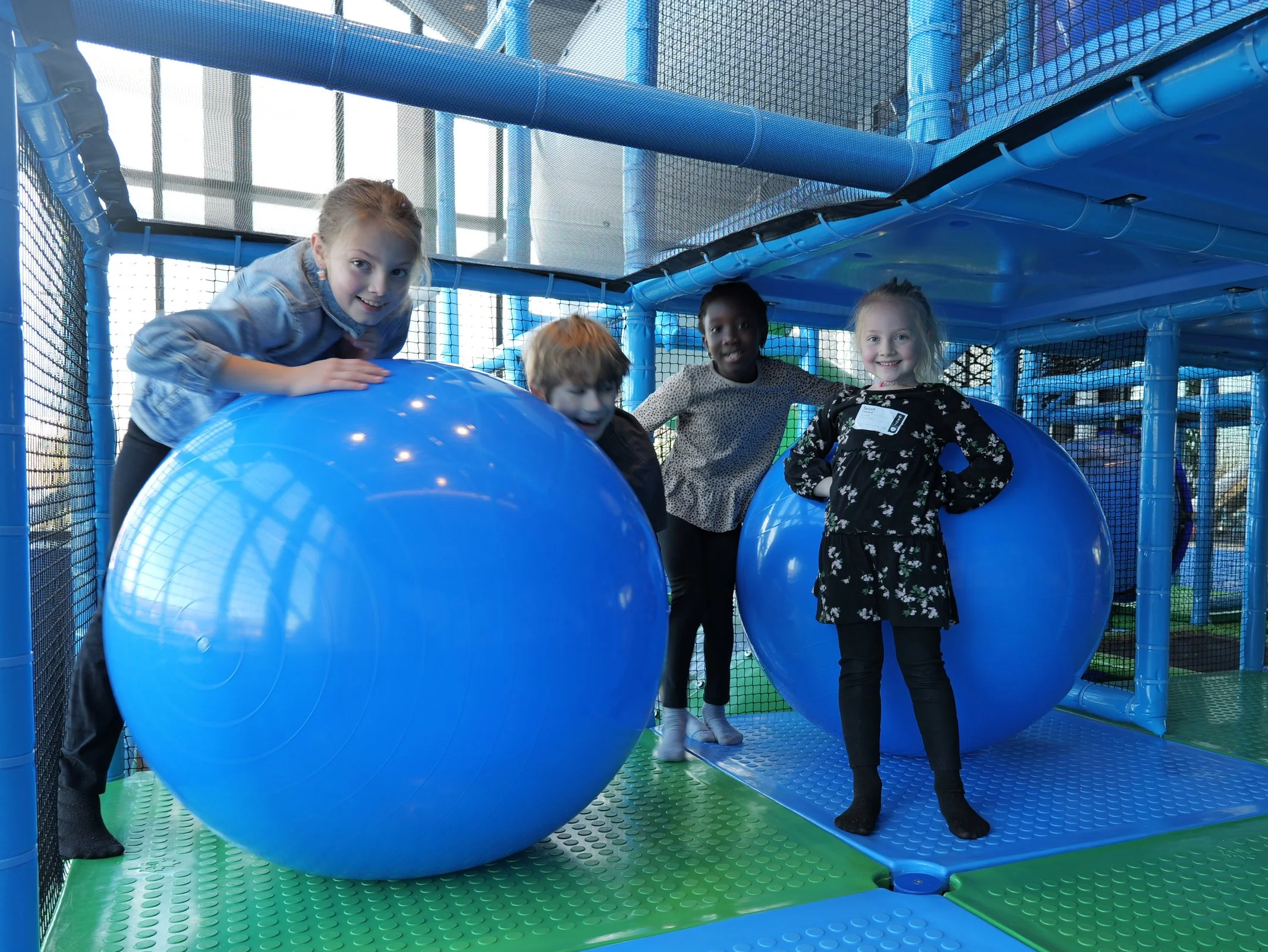 Four children playing inside an indoor play area with blue plastic climbing structures, a large blue ball, and green and blue floor mats at a church with a vibrant kids ministry.
