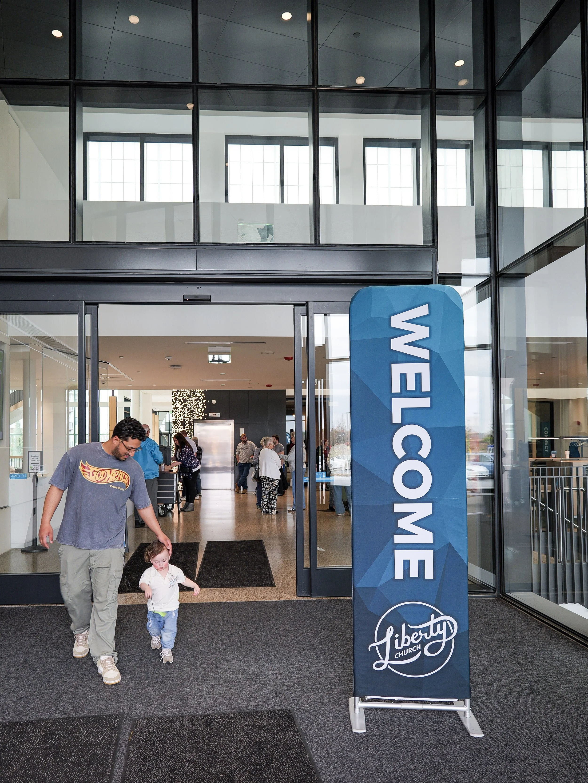 Entrance of a church with a sign that reads "liberty church" in front of the door. People, including a man and a child, walking out of the building. Glass windows and modern interior visible of a local church Woodbury.