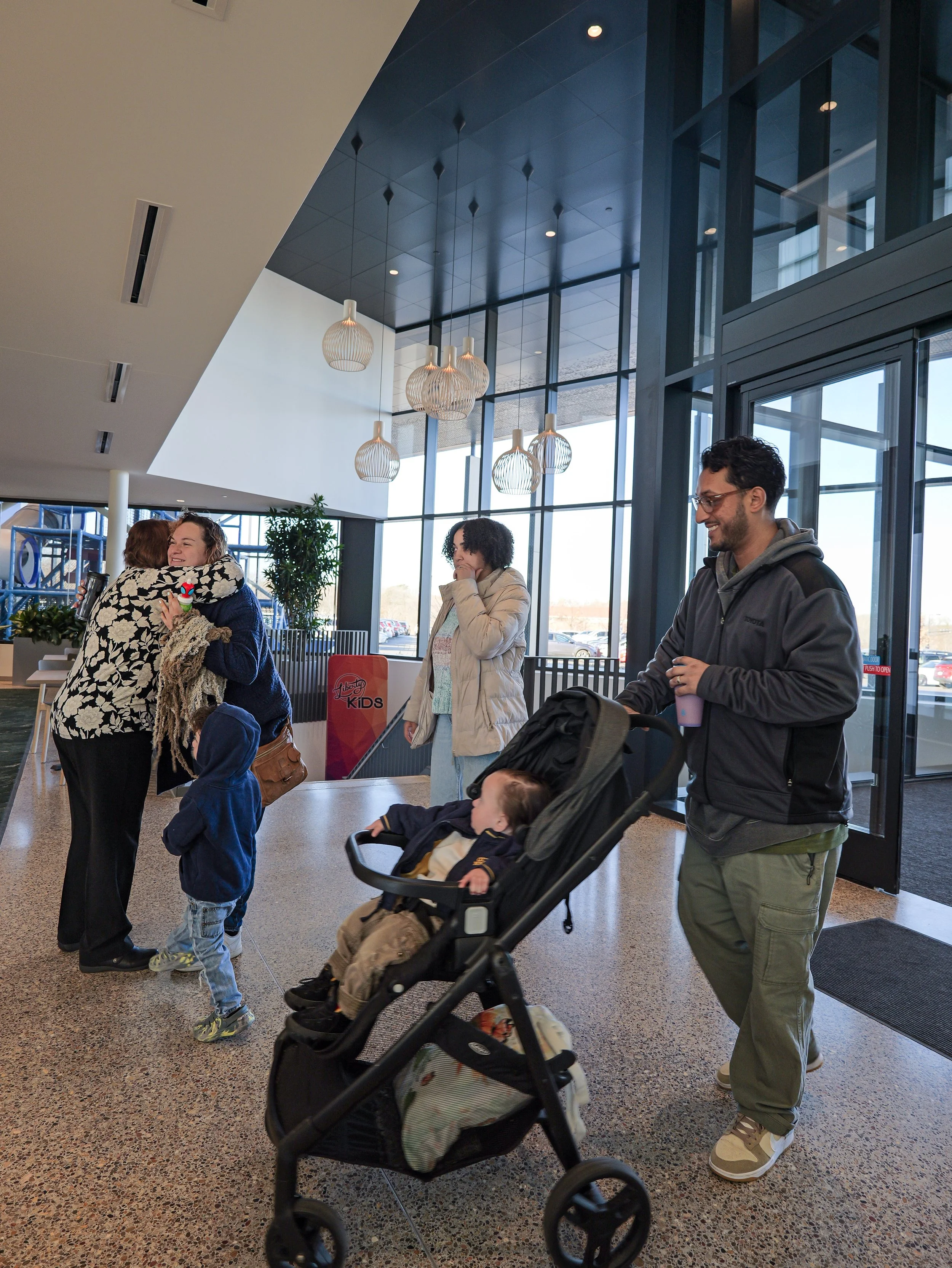 A group of people in a welcoming neighborhood church in Woodbury, with two women hugging, a young boy standing and looking at them, a woman with curly hair, and a man with glasses pushing a stroller with a child inside.