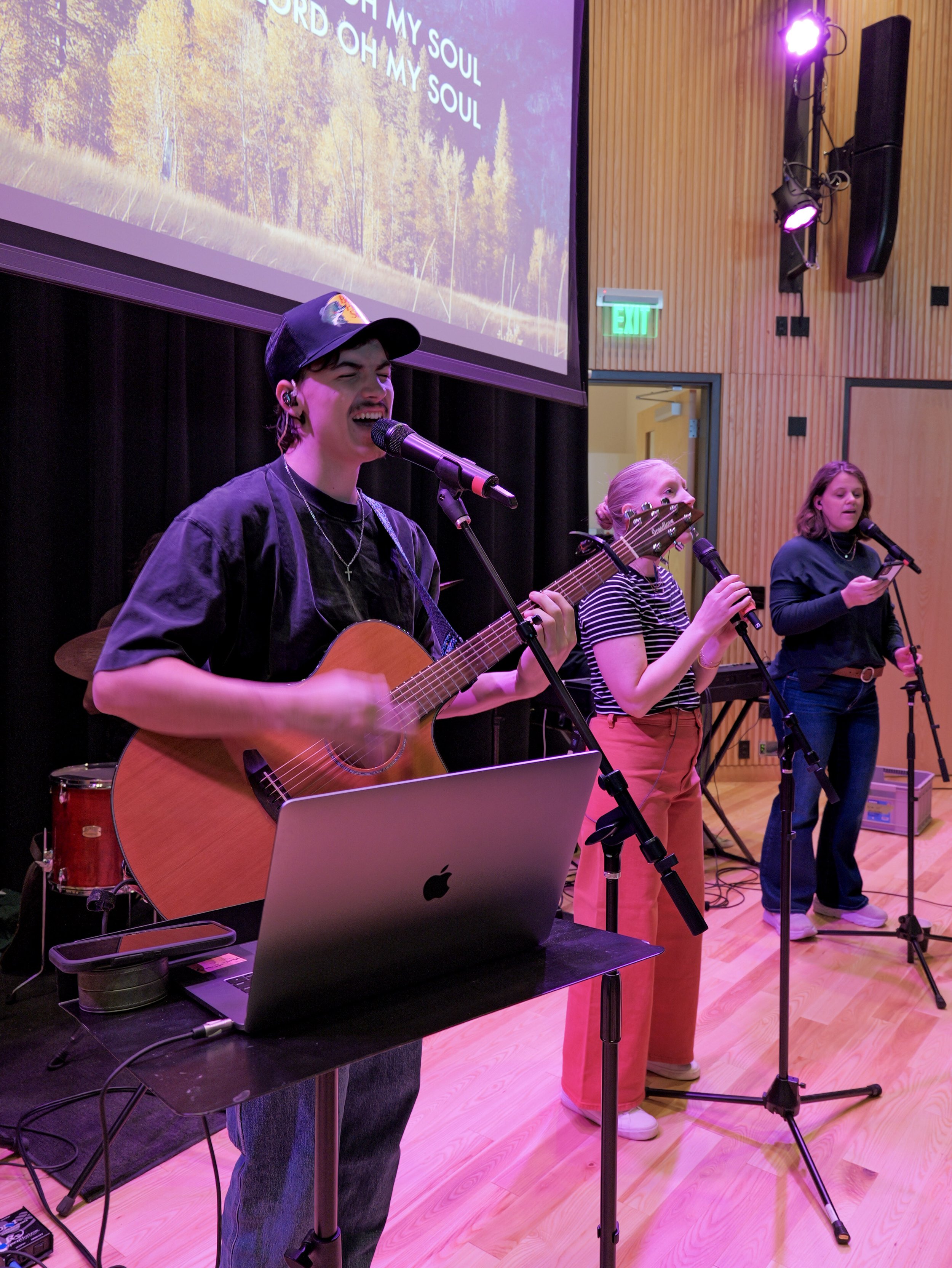 Three people singing and playing guitar on stage during a worship service in Woodbury, with a large screen behind them displaying song lyrics and a nature scene, in a room with wooden wall panels and stage lighting at a family church near me.