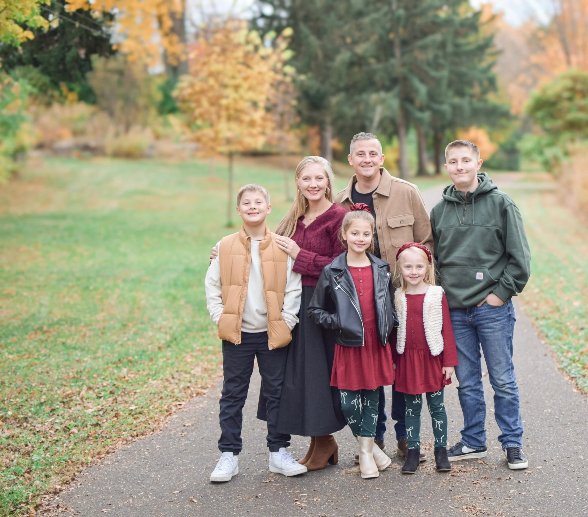 A welcoming pastor and his family of six standing outdoors on a fall day with colorful trees in the background.