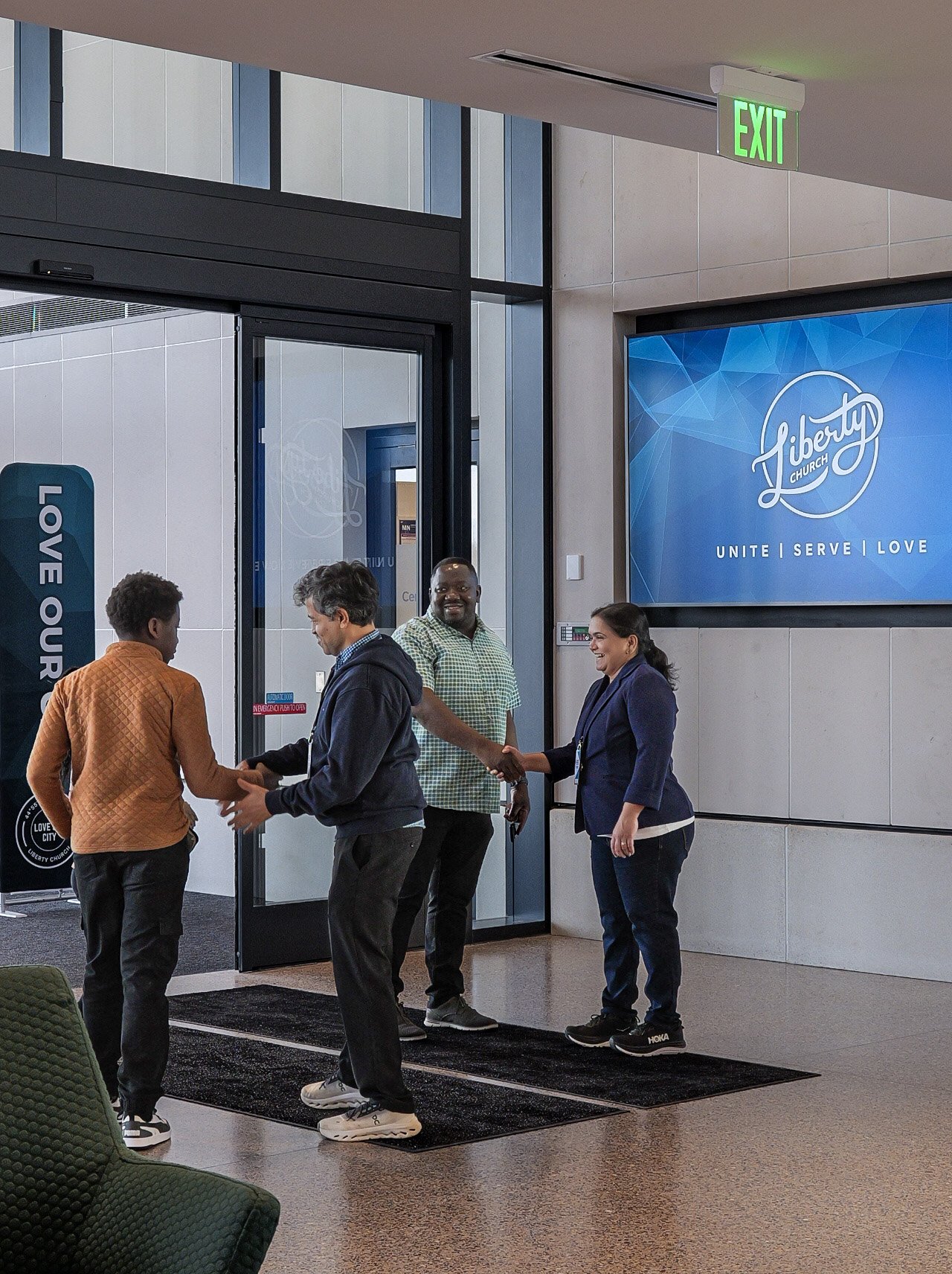A group of four people greeting and shaking hands in a lobby of Bible-based church Woodbury. The background features a large blue screen with the Liberty Church logo and the words "Unite | Serve | Love."
