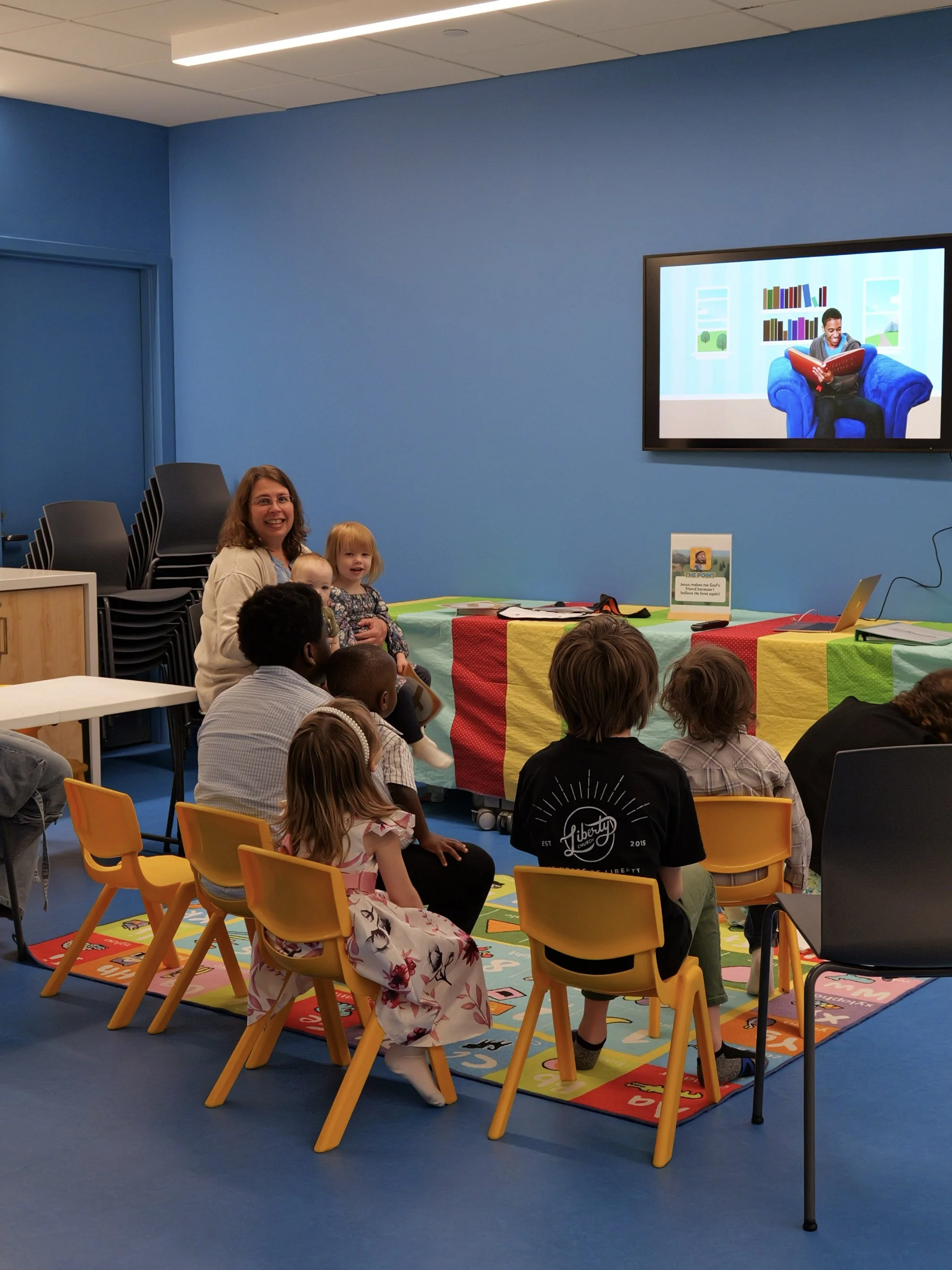 Children and adults sitting on yellow chairs and a colorful carpet in a room with a blue wall, watching a bible based curriculum on a large screen at a family centered church in Woodbury.