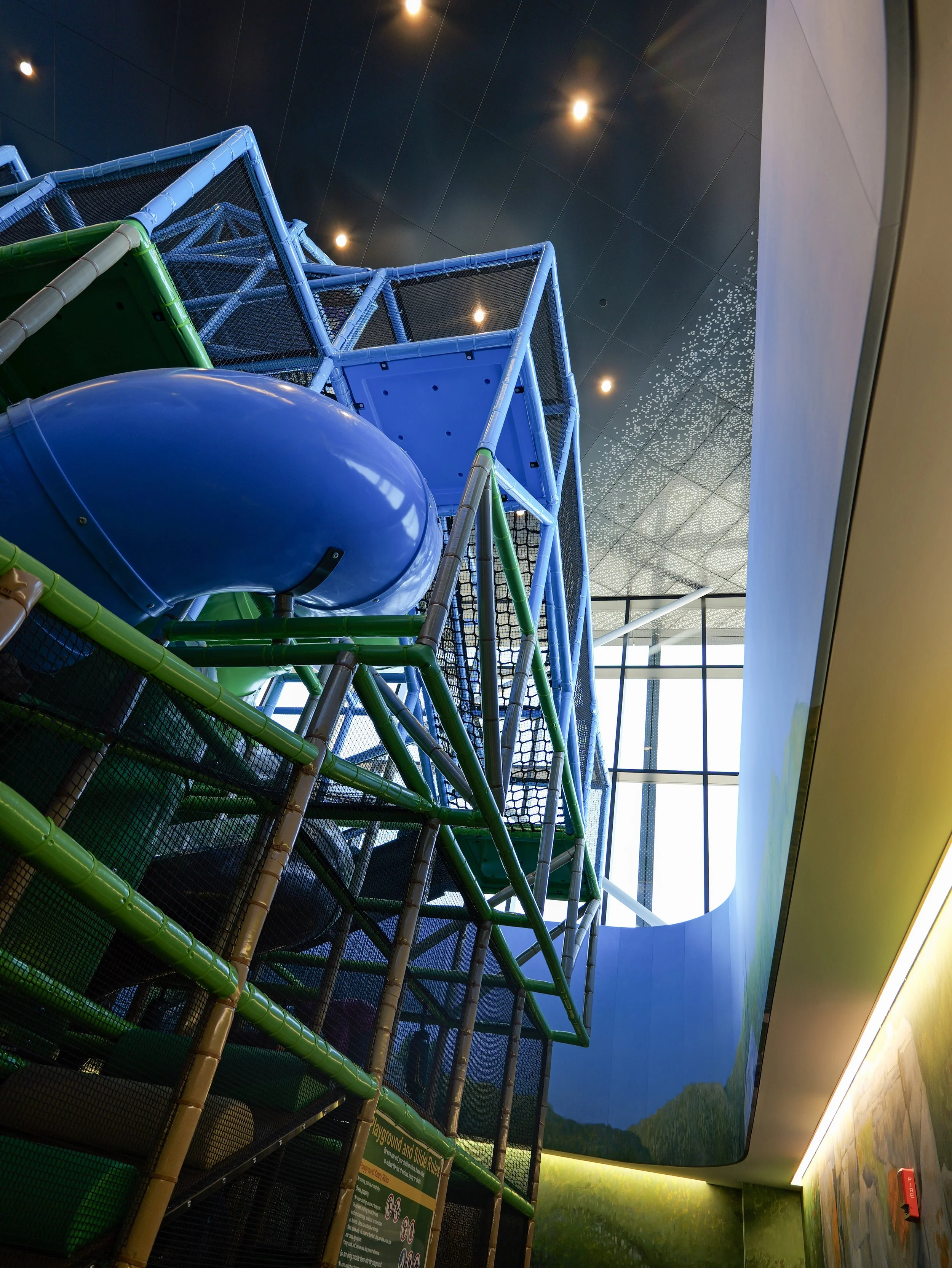 Indoor playground at a church for families in Woodbury Minnesota. Colorful climbing structures, green and blue tubes, and a slide, with a mural of mountains and trees on the wall and a dark ceiling with lights.
