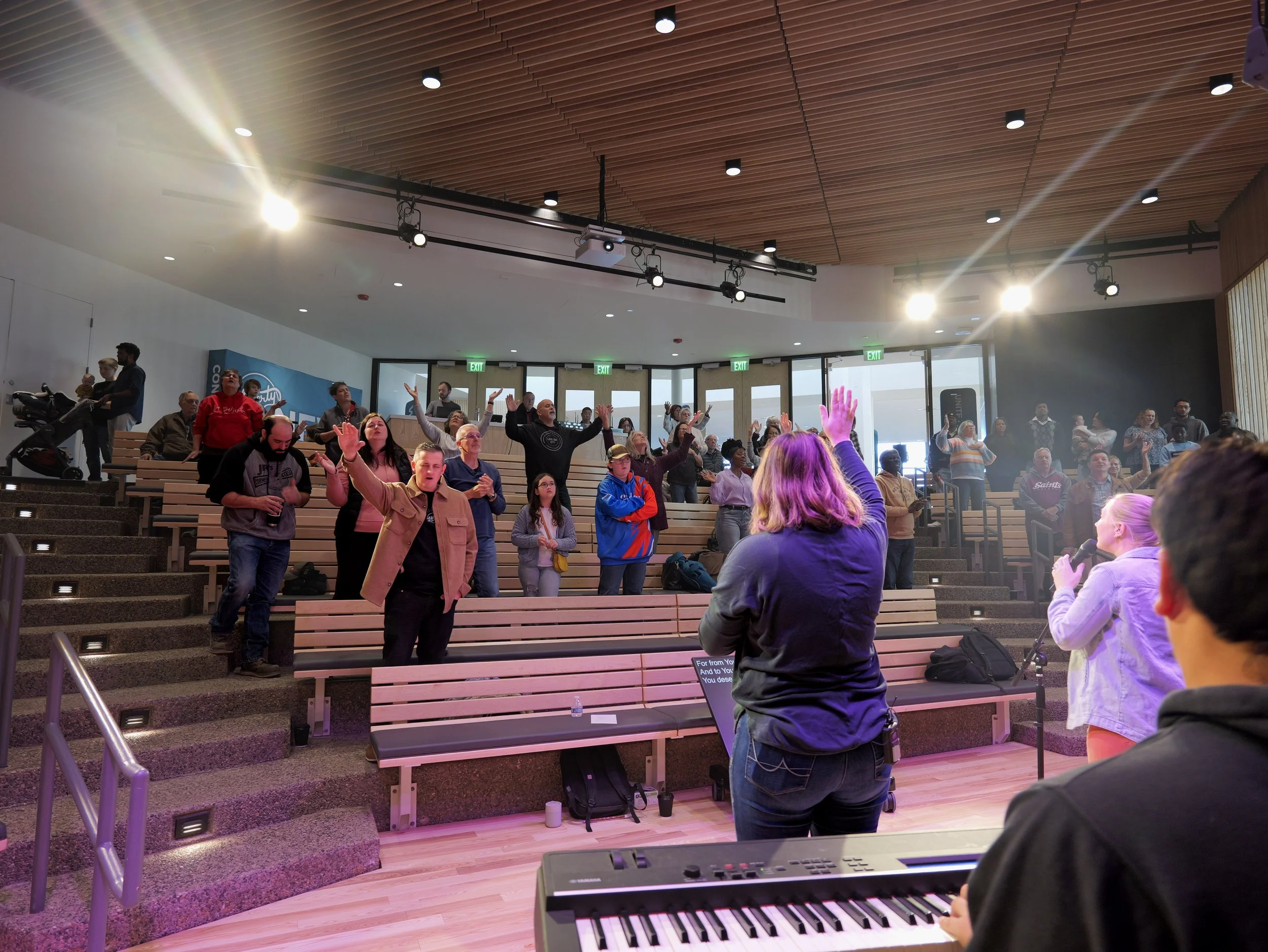 A diverse group of people on tiered wooden seating, some raising their hands and singing, in a modern auditorium with a wooden ceiling, stage lights, and large windows in the background.