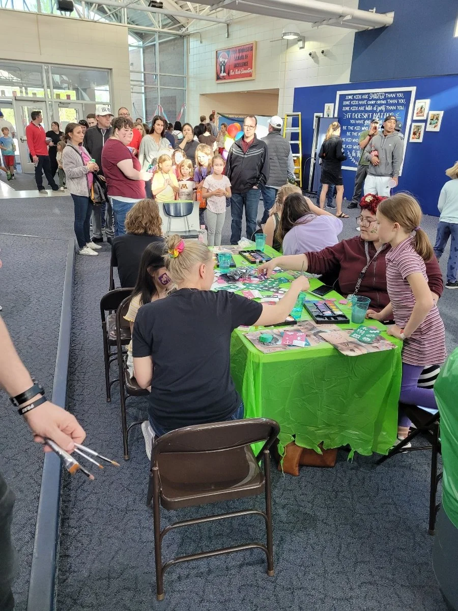 Children and adults gathered at an indoor event, with a table where face painting is being done and others waiting in line.