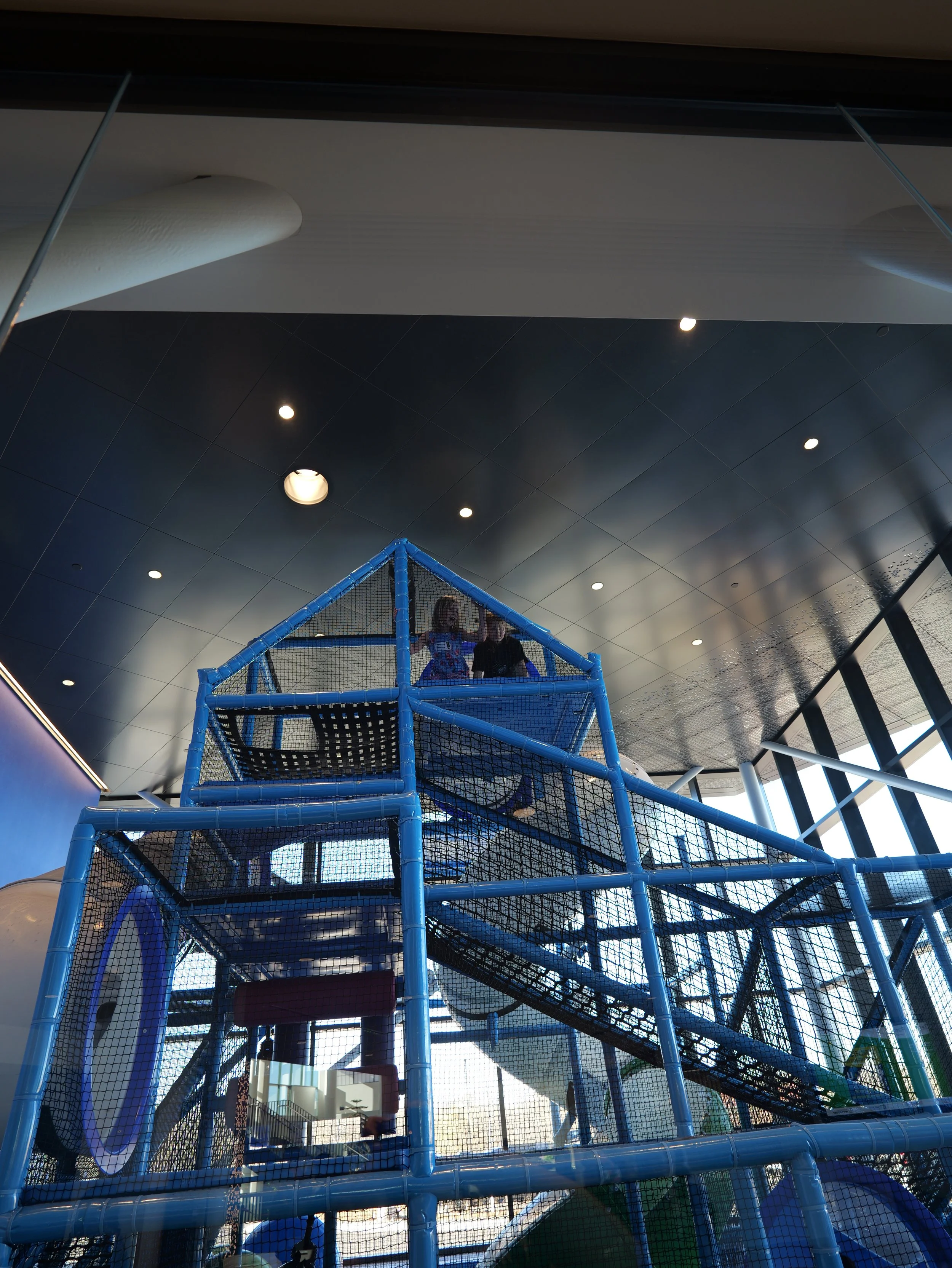 Indoor playground with a blue climbing structure, nets, slides, and children playing, located inside a modern building with large windows and ceiling lights.