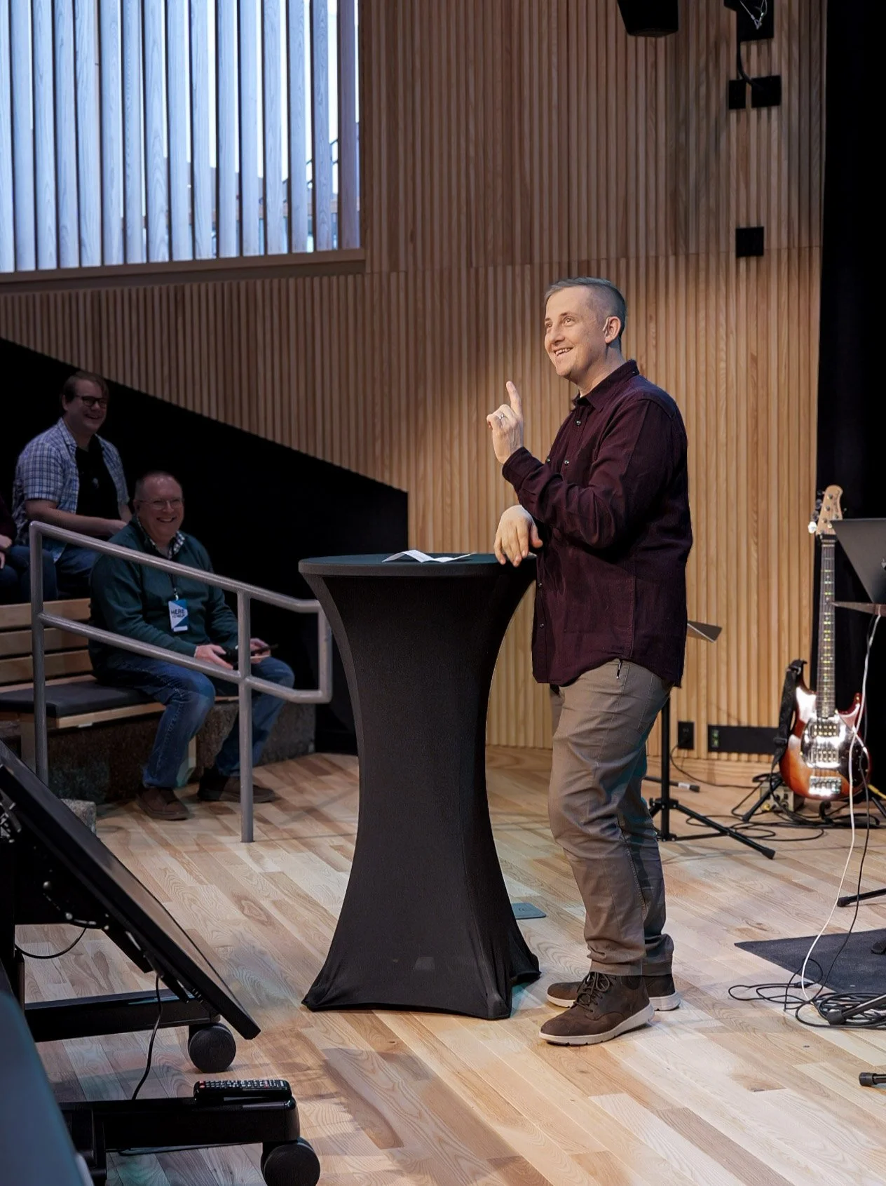 A man in a maroon shirt standing at a black lectern, speaking or presenting to an audience in a room with wooden paneling. Three people are sitting and smiling in the background, with one person on a staircase and another at a round table. Musical instruments and equipment are visible on the right side.