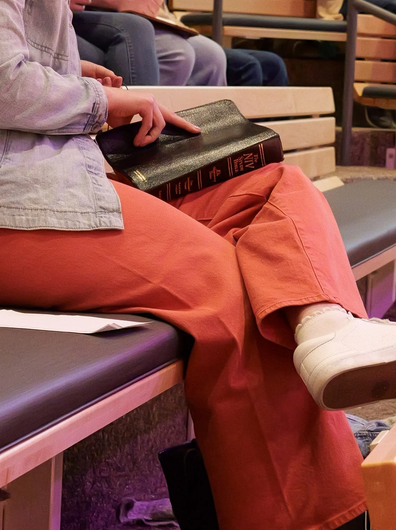 Person sitting cross-legged on a bench, holding a black book titled 'The New Sun' in their lap, wearing a light-colored denim jacket, coral-colored pants, white sneakers, and white socks, with other chairs and people visible in the background.