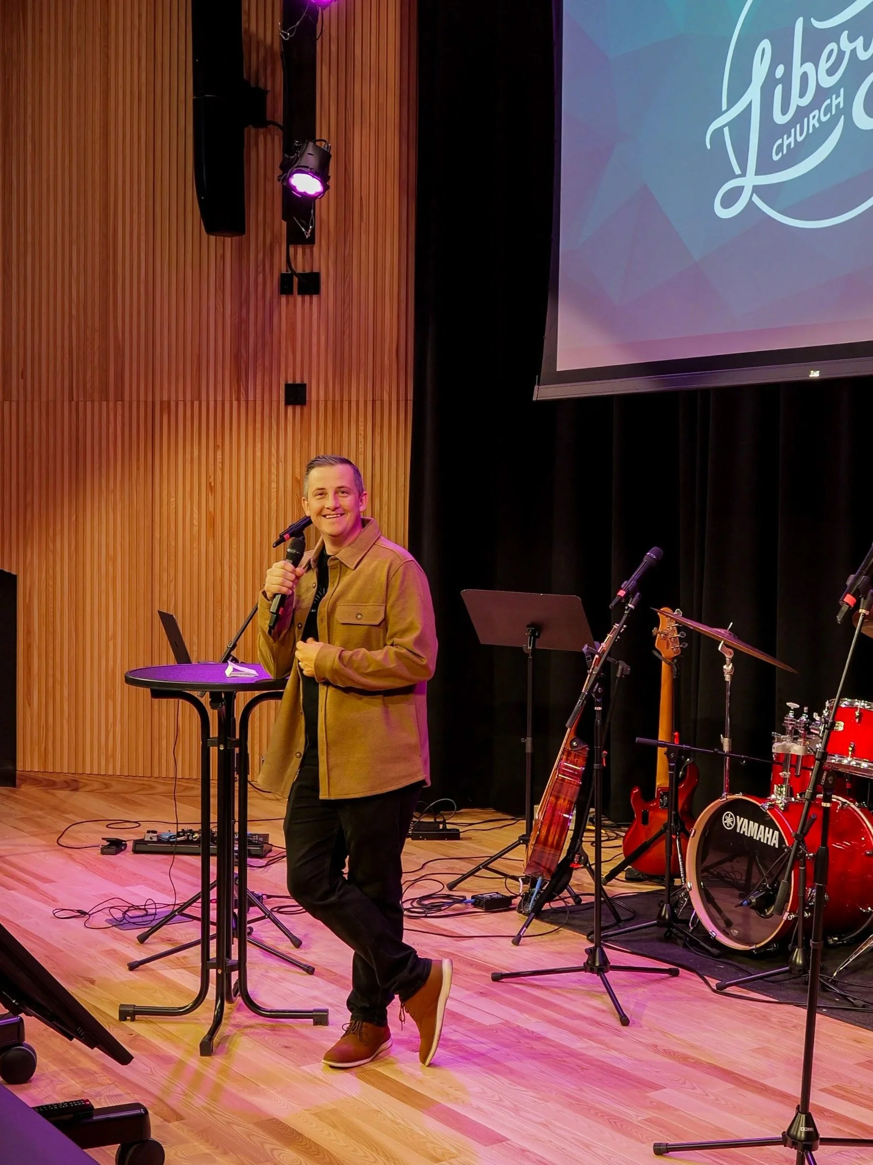 A pastor holding a microphone, smiling, standing on a stage at a Woodbury church. Wooden wall background, stage lighting, and a large screen displaying a logo for Liberty Church.