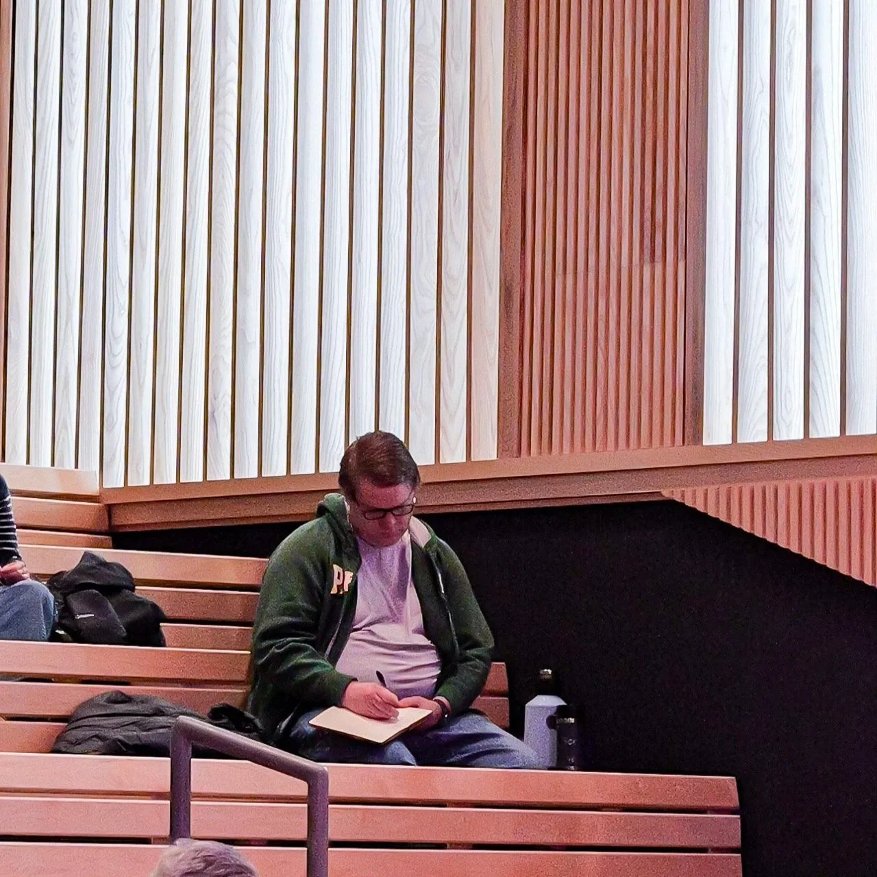 A young man with glasses, wearing a green hoodie and white T-shirt, sitting on wooden bleachers and writing in a notebook. There are backpacks and water bottles nearby, and a wood-paneled wall in the background.