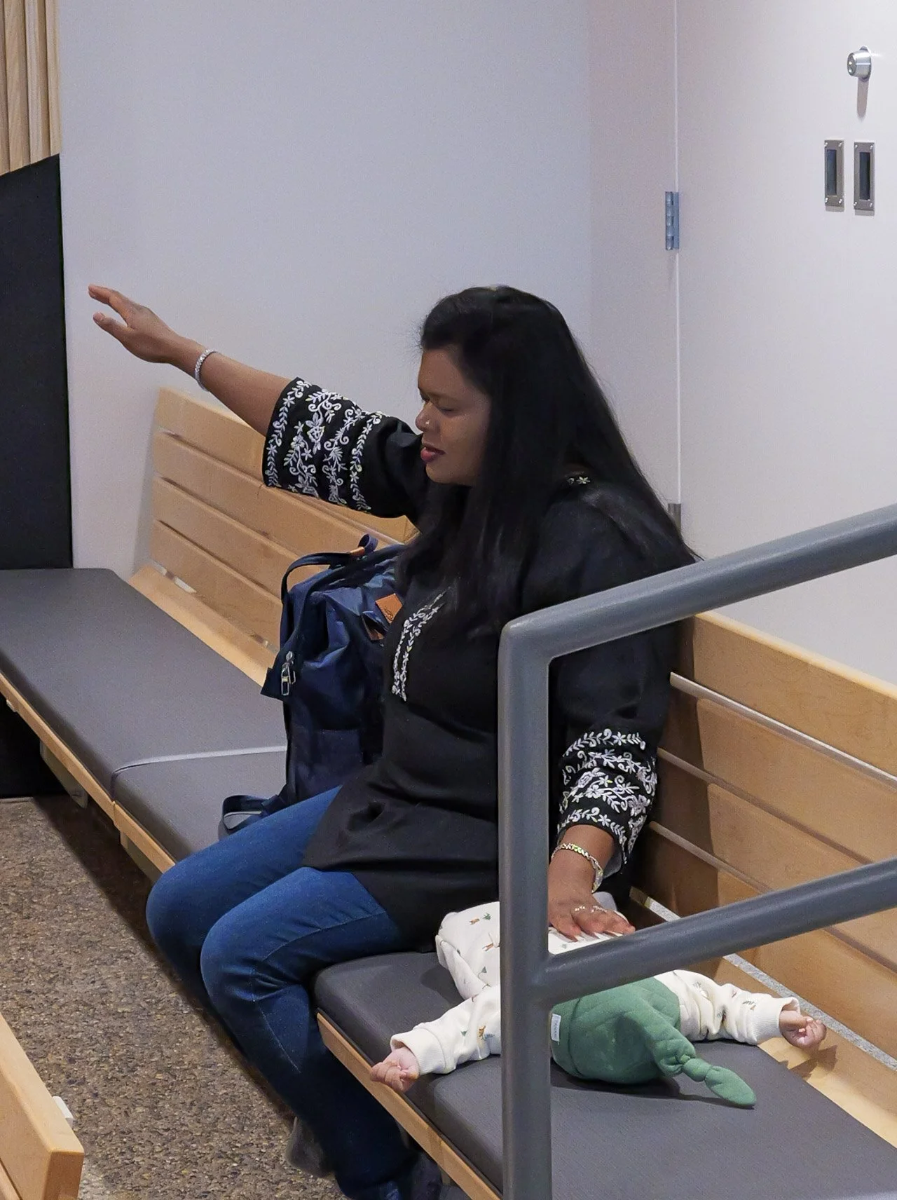 A woman with long dark hair and a black shirt with white embroidery on the sleeves sits on a bench with her eyes closed and her arm extended, appearing to be praying or meditating. A small baby dressed in white and green lies on the bench next to her, reaching out with arms extended. The setting appears to be indoors, possibly in a waiting area.