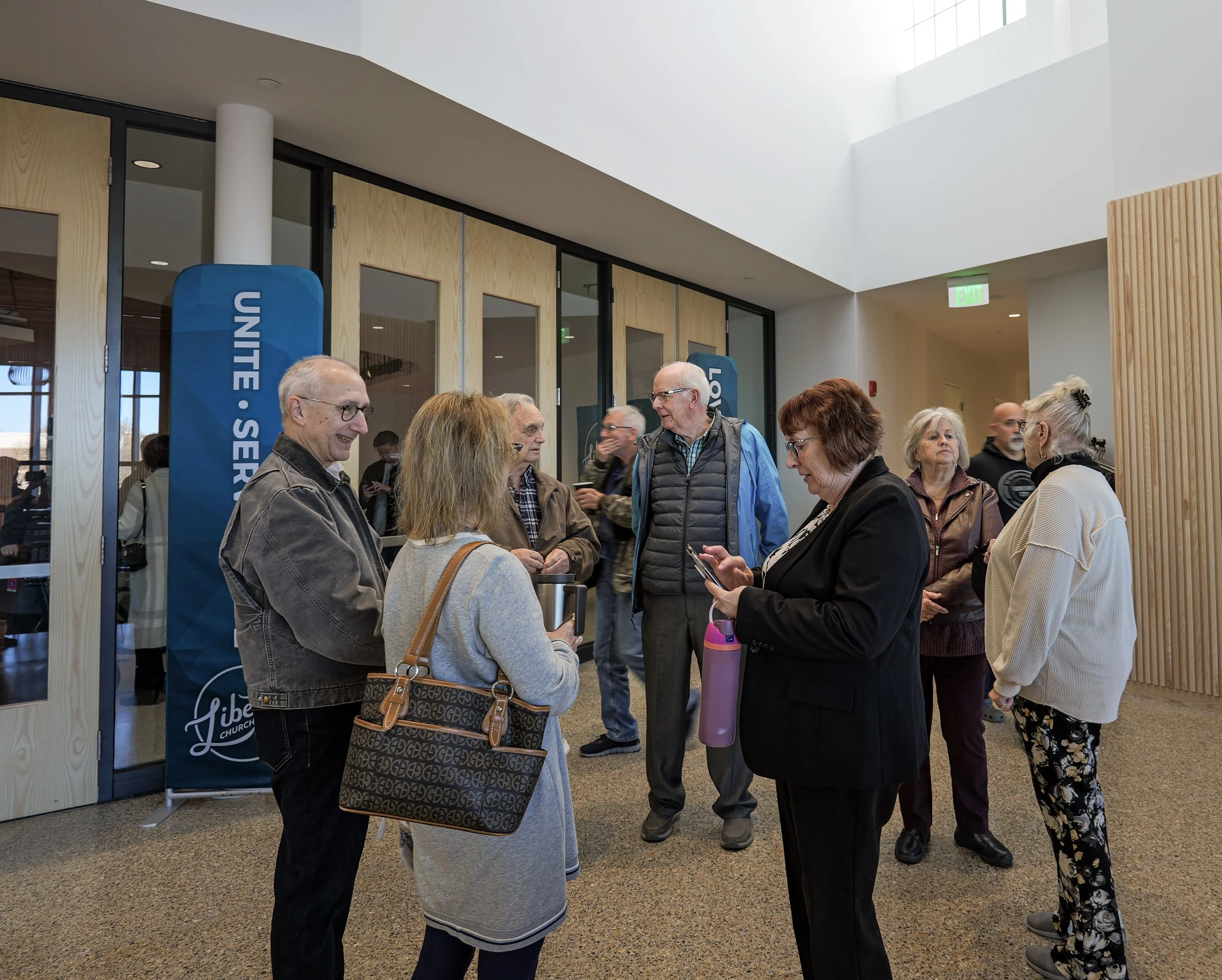 Group of people socializing indoors near a draped partition with a sign that reads 'UNITE SERVE LOVE' at the entrance to an Assembly of God Church. 