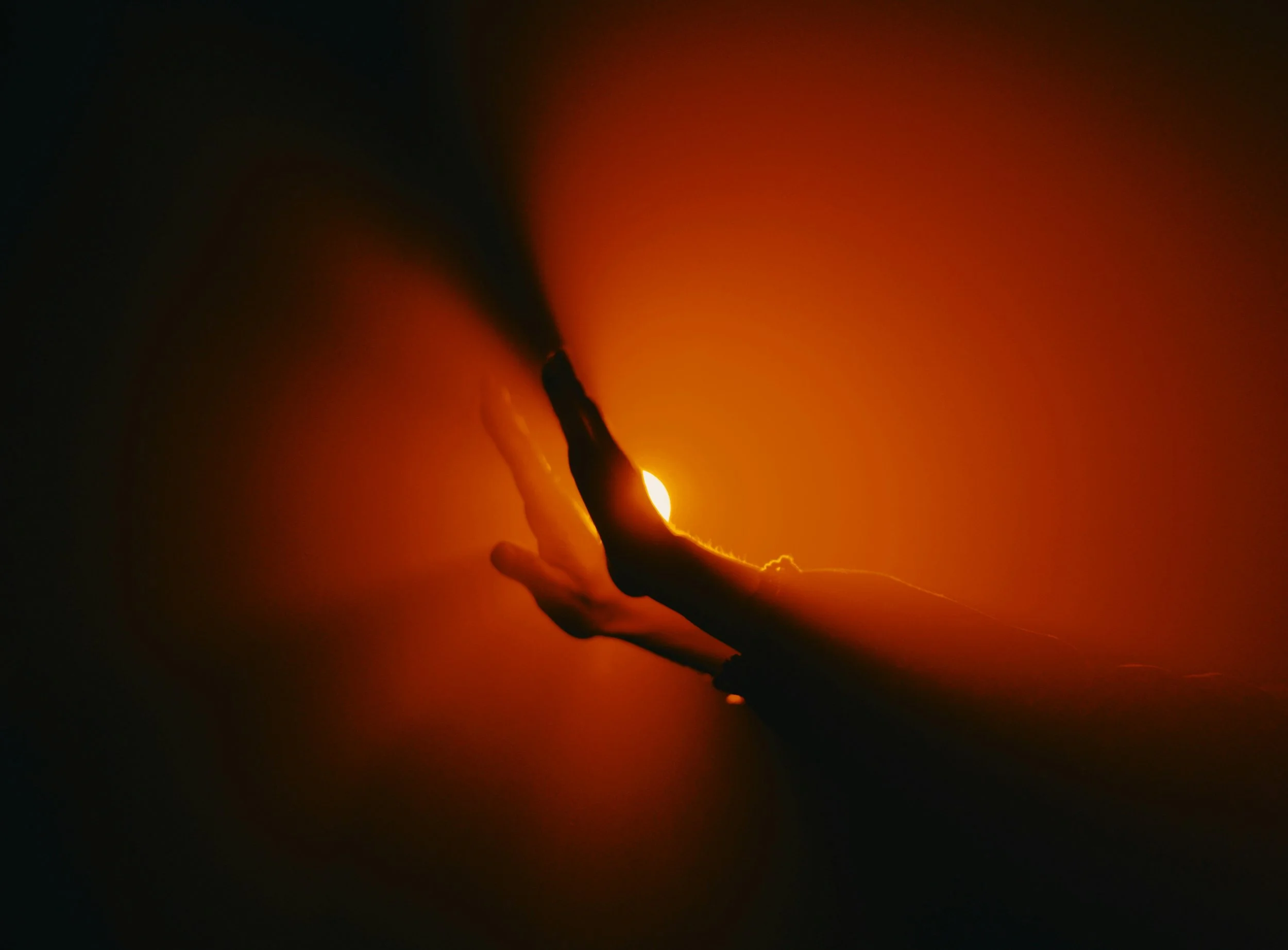 A person's hand holding a lit matchstick with the flame burning brightly, casting an orange glow in a dark environment.