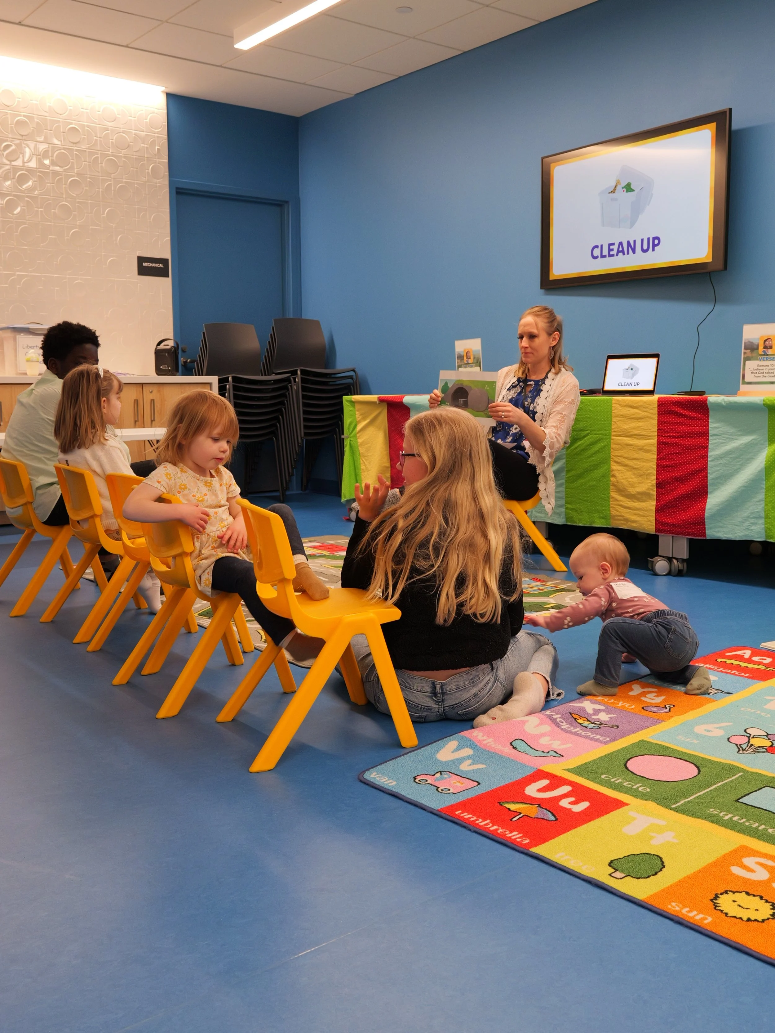 A classroom with children sitting on yellow chairs and listening to a woman reading a bible based picture book. A colorful educational rug is on the blue floor, at Liberty church's kids ministry.