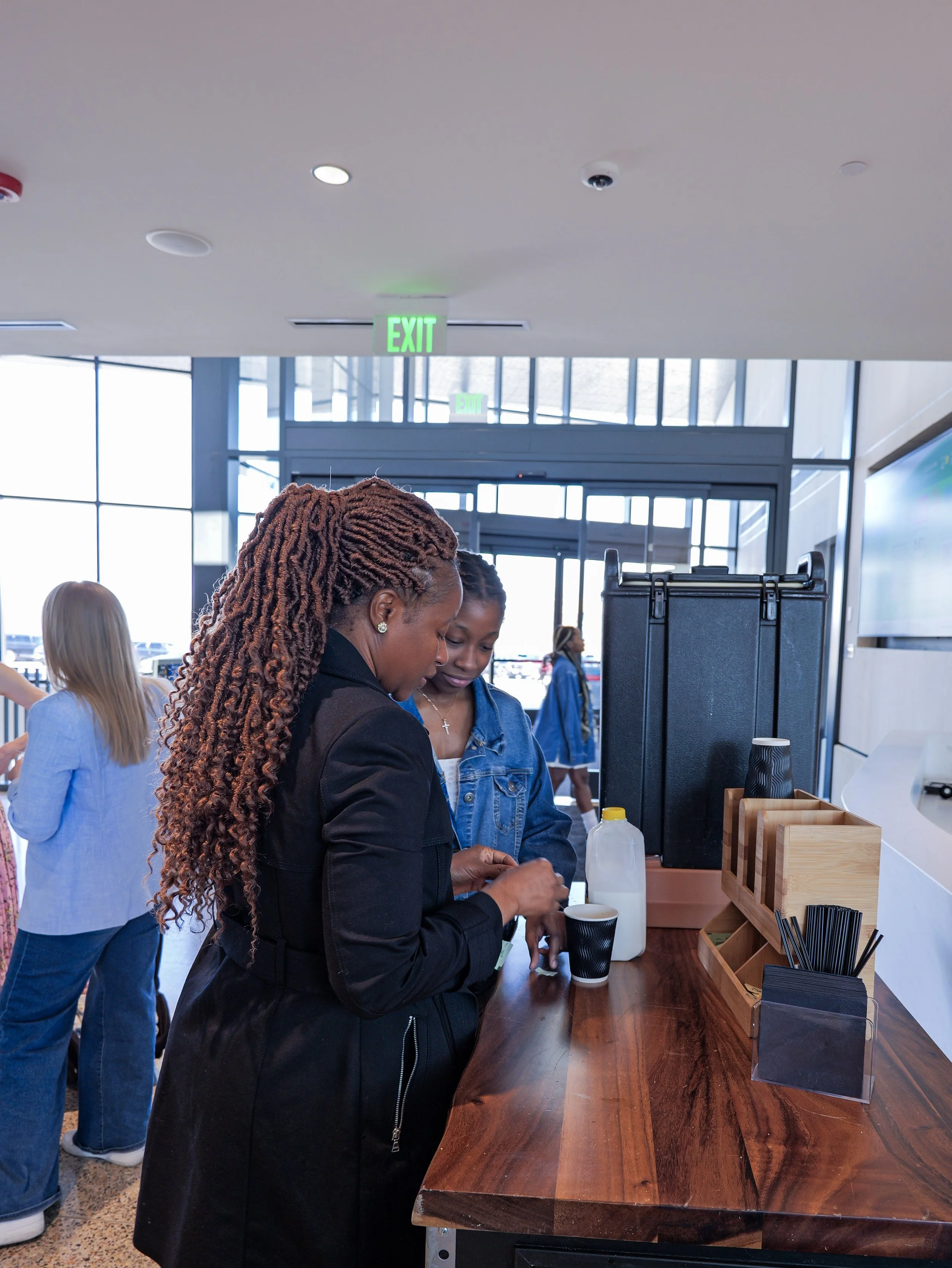 People at a wooden counter in a modern indoor space with large windows. A woman with reddish-brown curly hair is filling a cup, and other people are visible in the background. An exit sign is seen overhead.