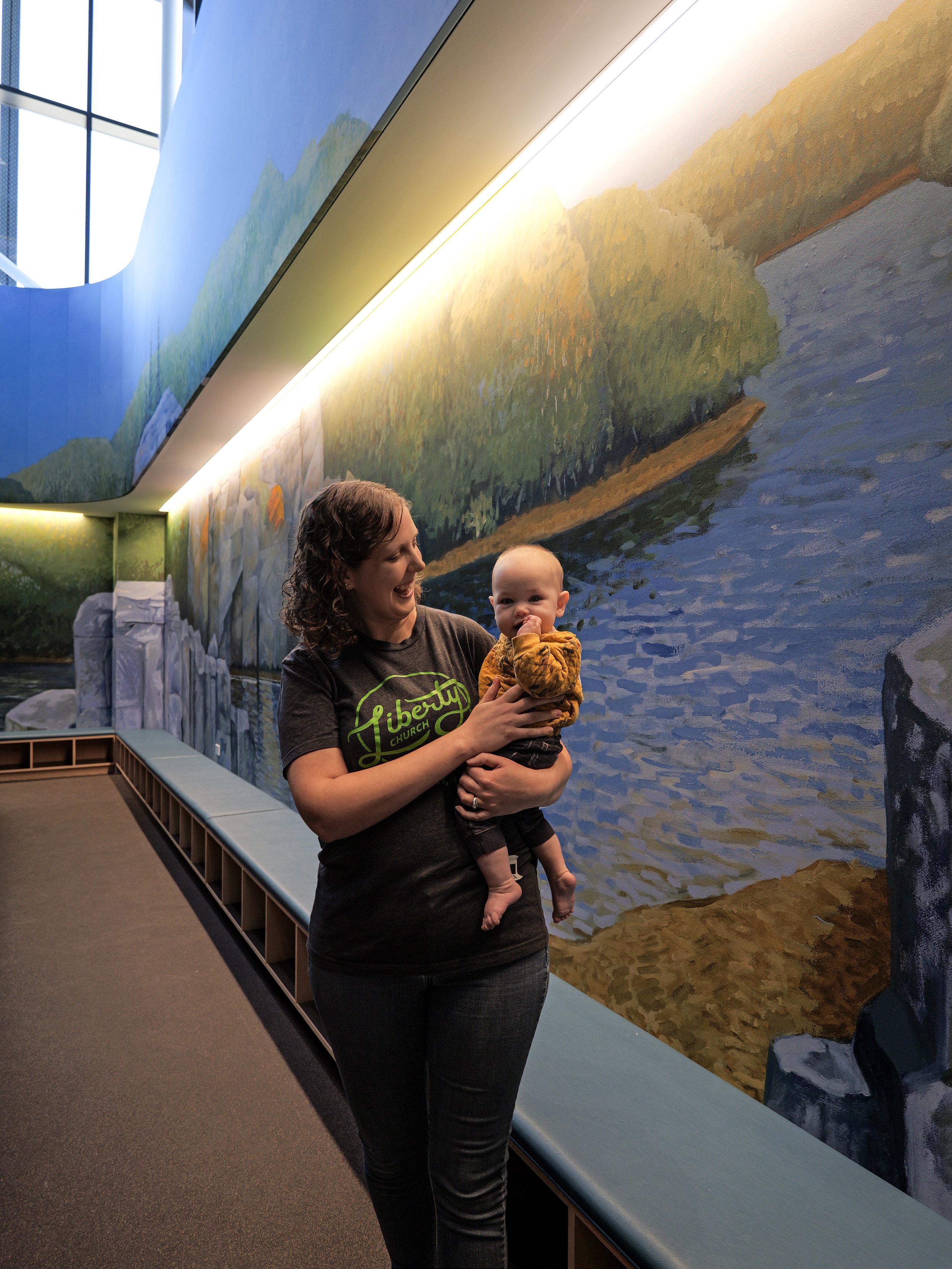 A woman holding a baby in front of a large mural painting of a river, trees, and sky, at a church with a thriving children's ministry..