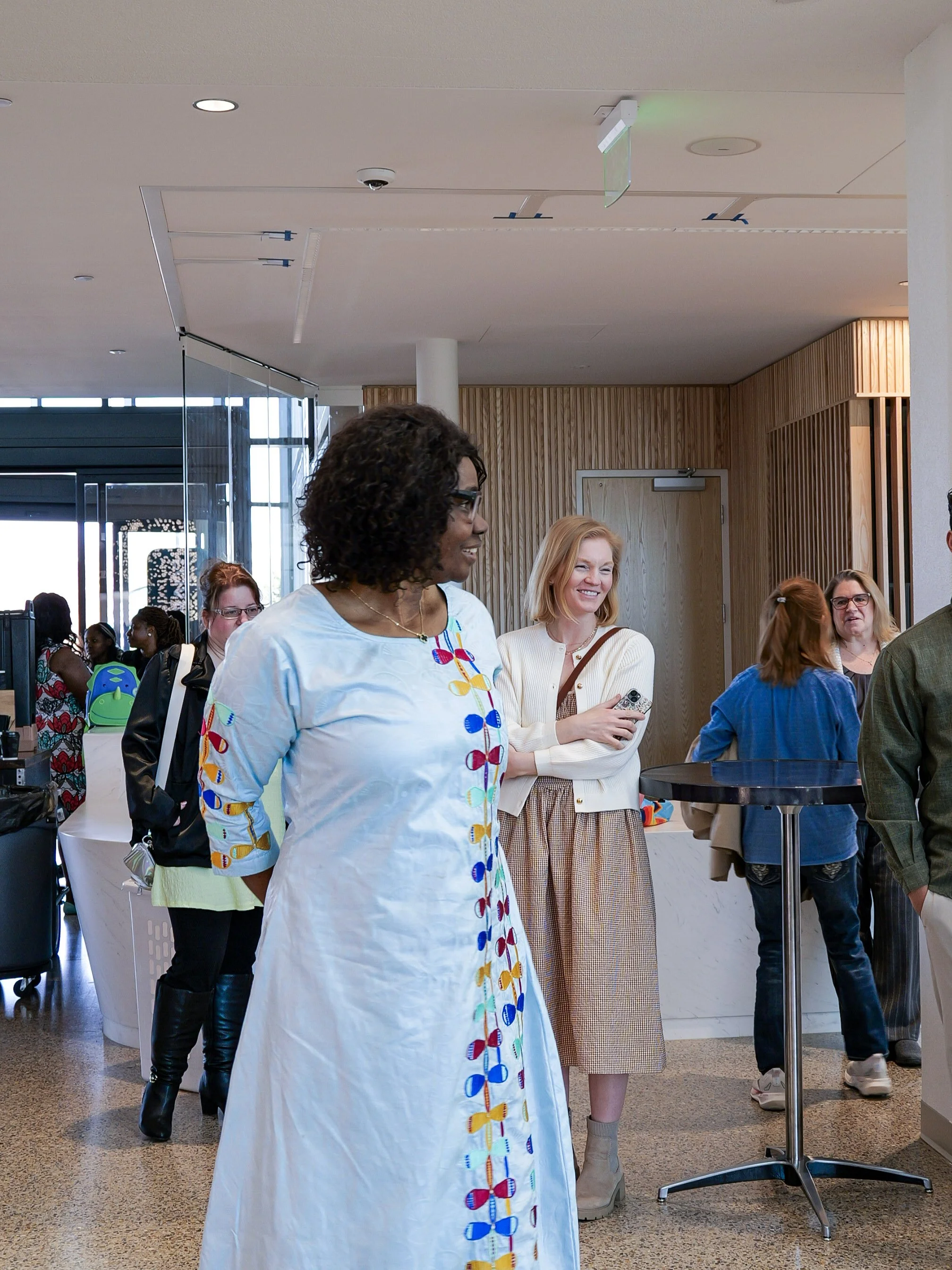 A diverse group of people gathered indoors at a church for families, including a woman in a white dress with colorful bow embroidery, smiling and engaged in conversation. 
