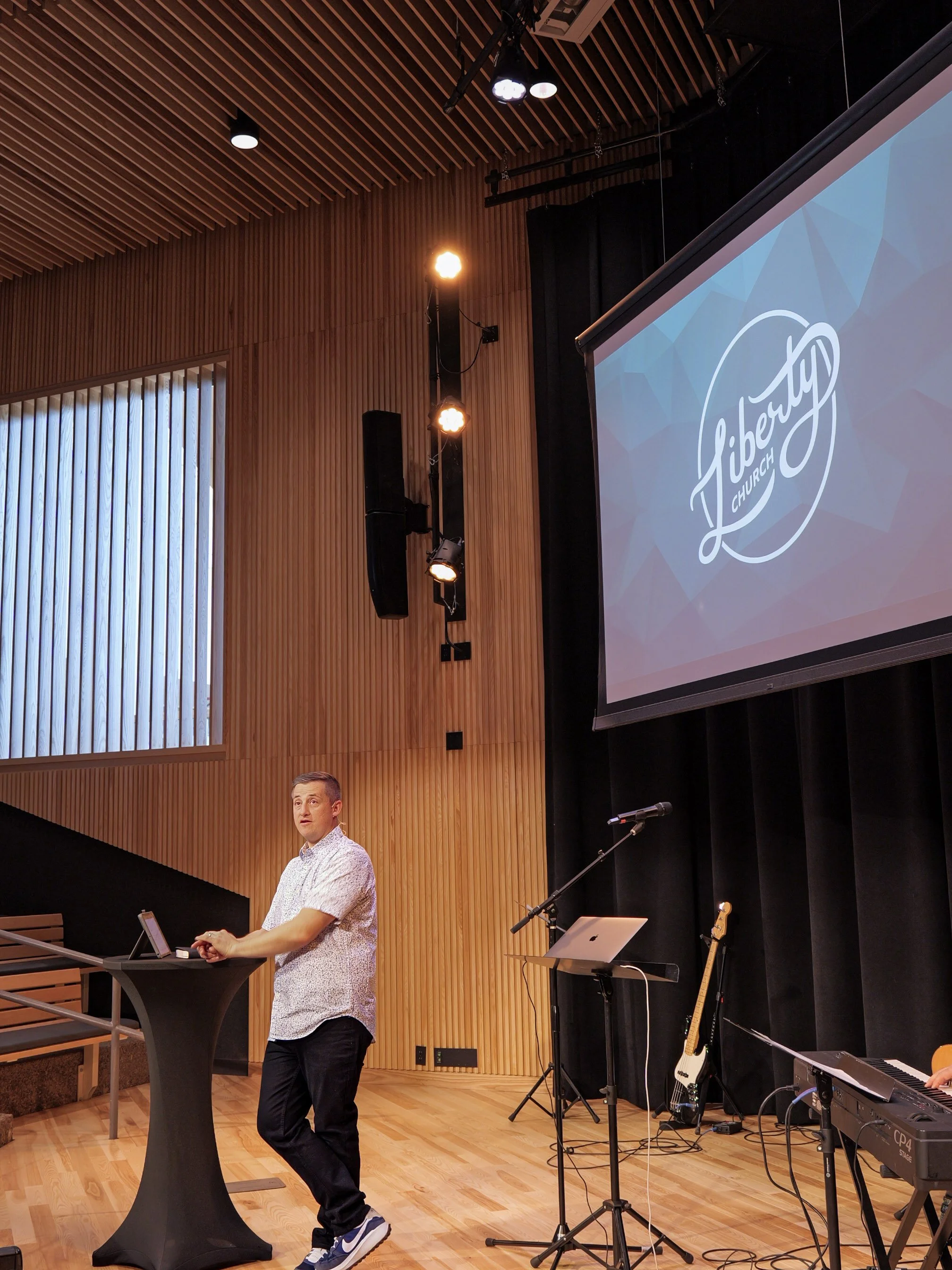 A man stands on a stage with musical instruments nearby, including a guitar and keyboard. He appears to be speaking or presenting, with a presentation slide displaying 'Liberty Church' logo projected behind him. The room has wooden walls and ceiling panels, with stage lighting focused on him.
