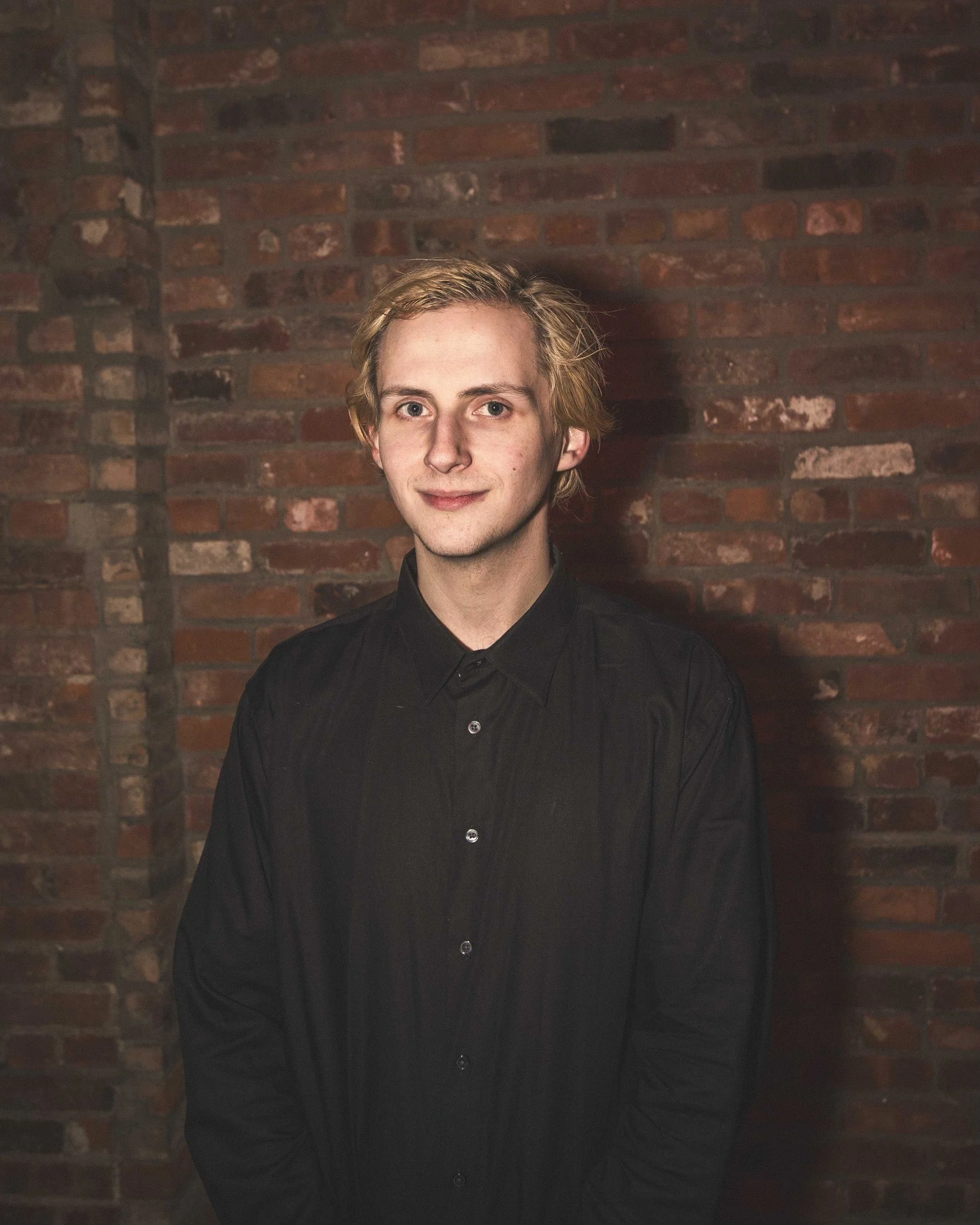A young man with light blonde hair, fair skin, and blue eyes, wearing a black button-up shirt, standing against a red brick wall background.