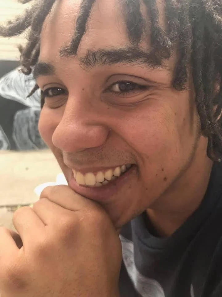 Close-up of a smiling young man with dreadlocks, resting his chin on his hand, in an indoor setting.