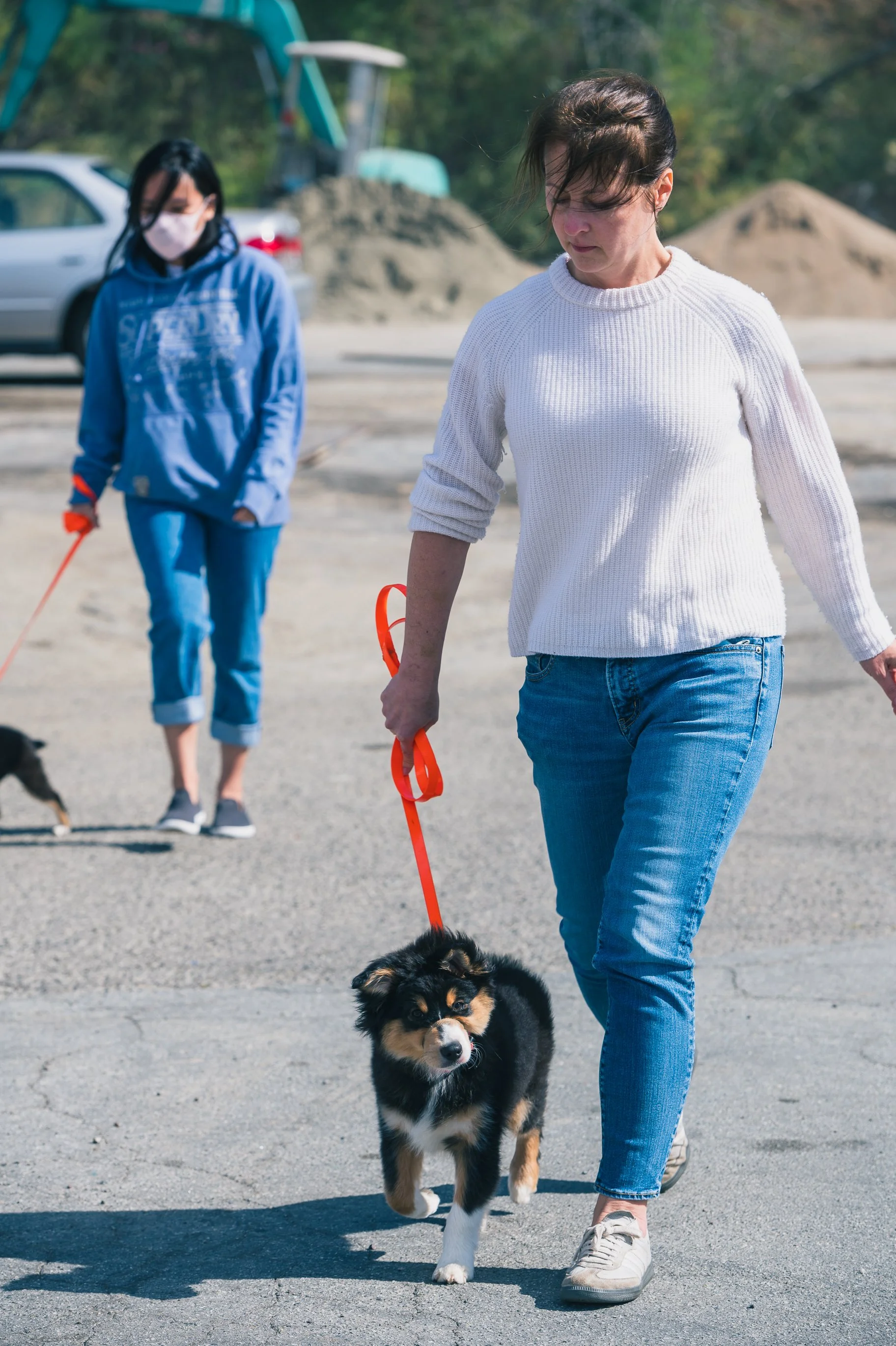 A woman walking a puppy on a leash outdoors, with another woman in the background and construction materials nearby.