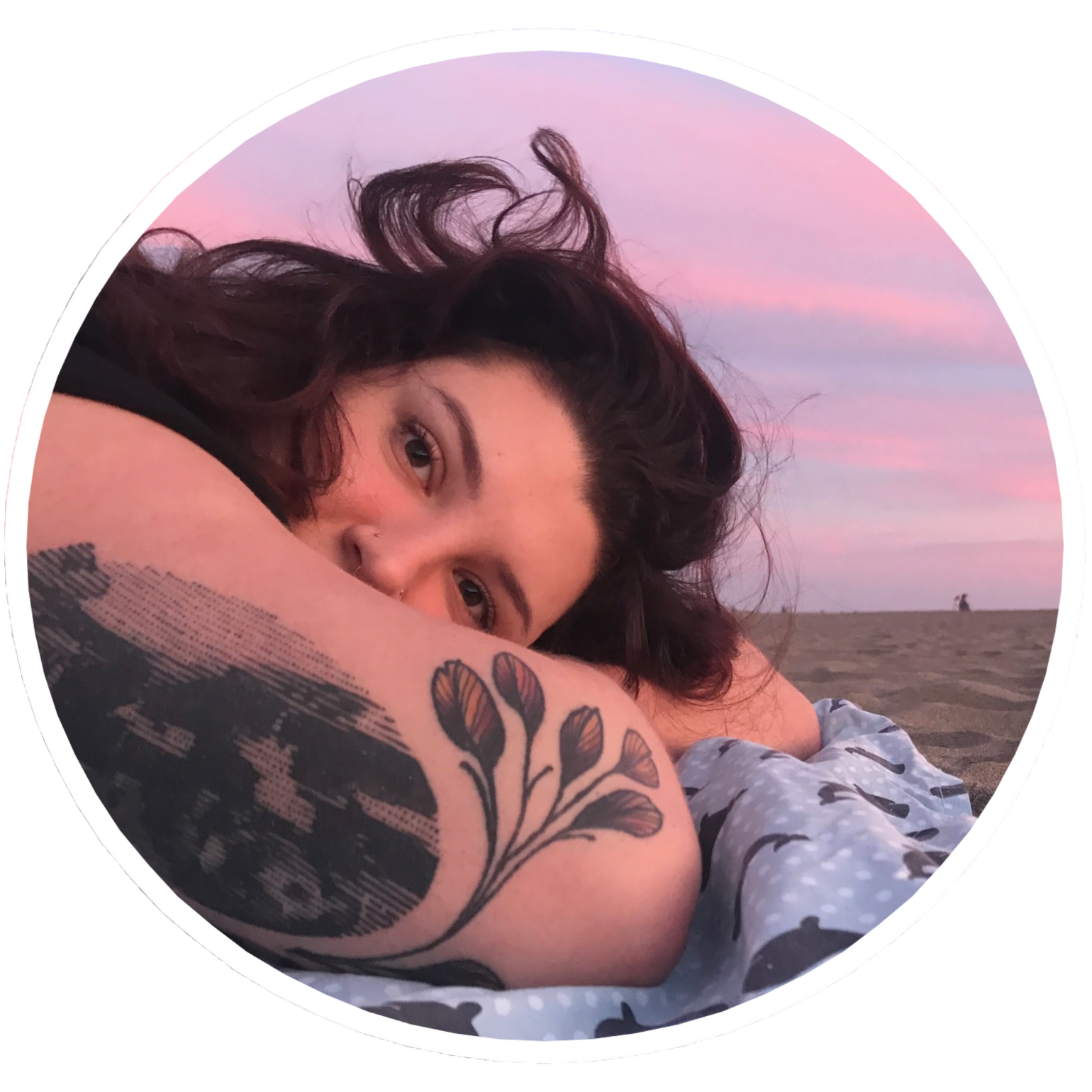 A woman with dark curly hair lying on her front on a beach during sunset, resting her head on her arm with a tattoo of flowers on her forearm, looking at the camera with a soft expression, beach and pink sky in the background.