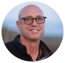 A man with a bald head and glasses smiling outdoors, wearing a dark jacket.