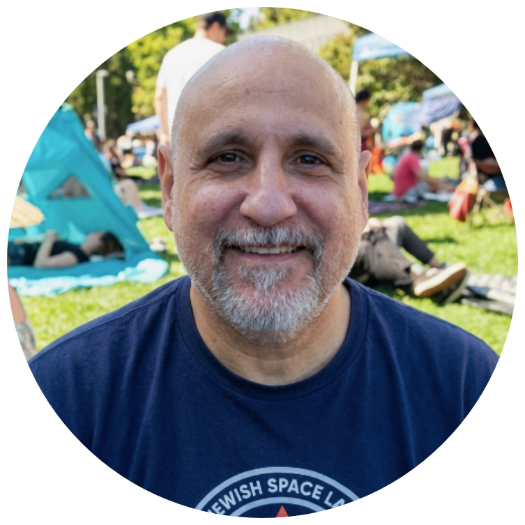 Smiling middle-aged man with a beard and mustache sitting outdoors at a gathering or festival, with tents and people in the background.