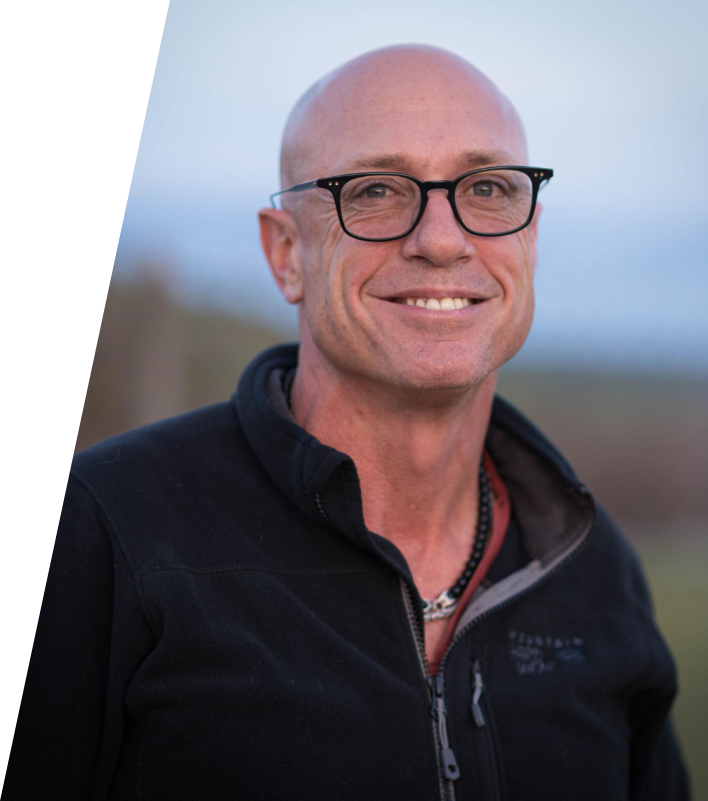 A smiling middle-aged man with glasses, wearing a black zip-up jacket outdoors during daytime.