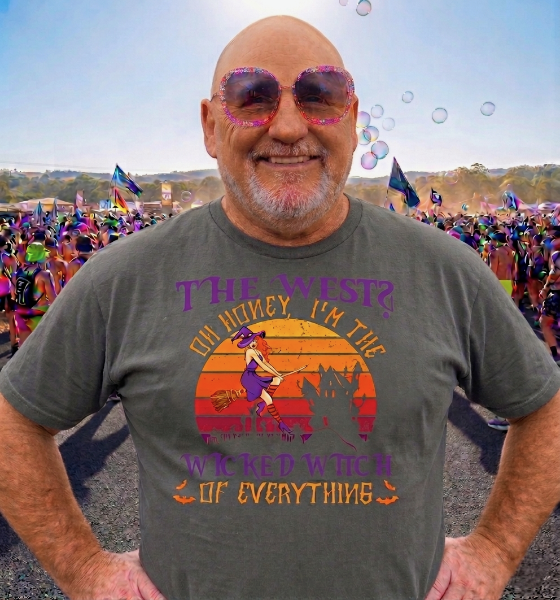 A man with a bald head and sunglasses smiling at a pride parade, wearing a graphic t-shirt with a witch and sunset design and colorful text referencing the Wizard of Oz.