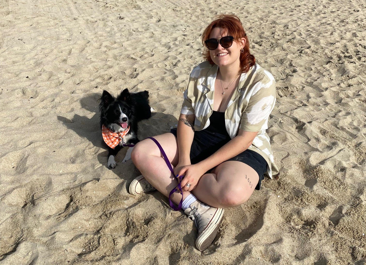 A woman with red hair and sunglasses sitting cross-legged on the sand next to a black and white Border Collie dog wearing an orange checkered bandana at the beach.