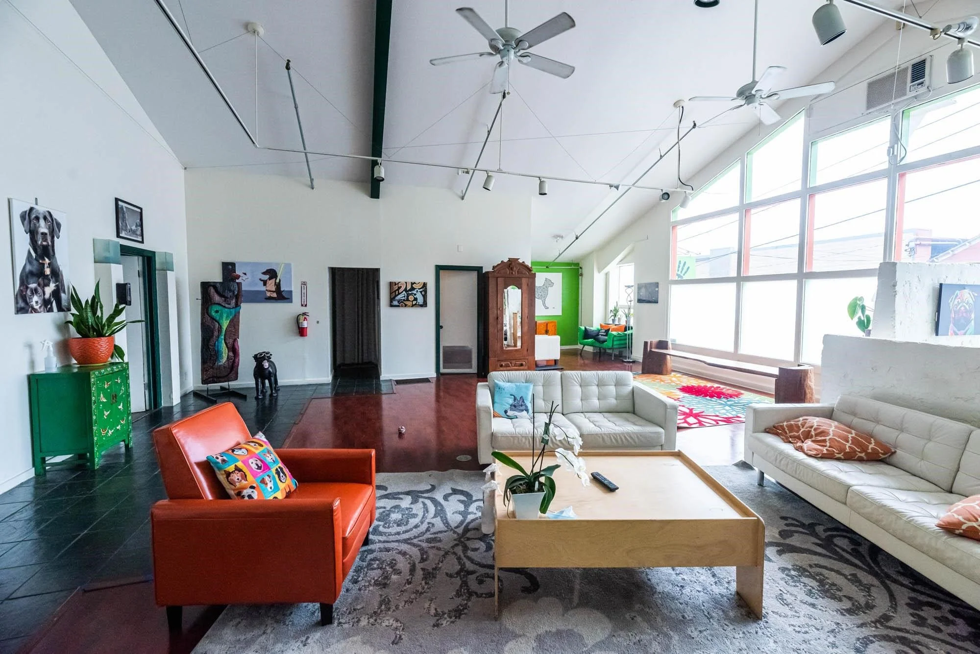 A bright, spacious living room with high, sloped ceiling and large windows. The room features white and green walls with several colorful paintings and artwork. There is a white sofa with a Giraffe pillow, a red armchair with a cartoon-themed pillow, and a wooden coffee table. Various houseplants and modern decorations are also present, along with a beige patterned rug on dark flooring. Ceiling fans and track lighting illuminate the space.